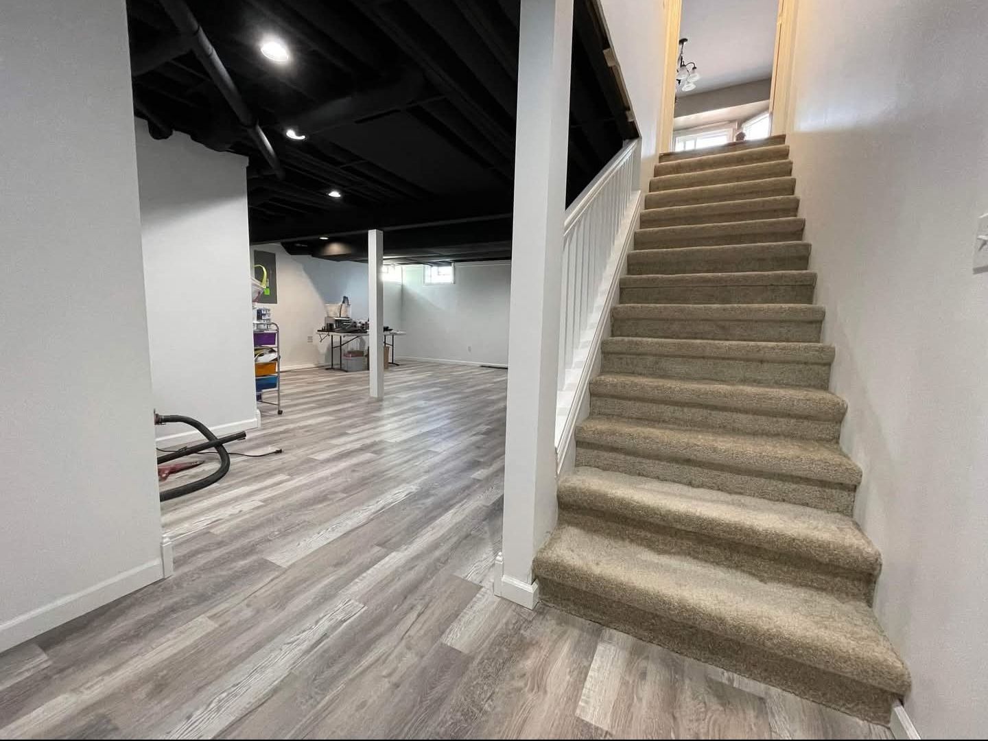 Staircase leading up with carpeted steps. Basement with wood-look flooring and black ceiling, workout equipment visible.