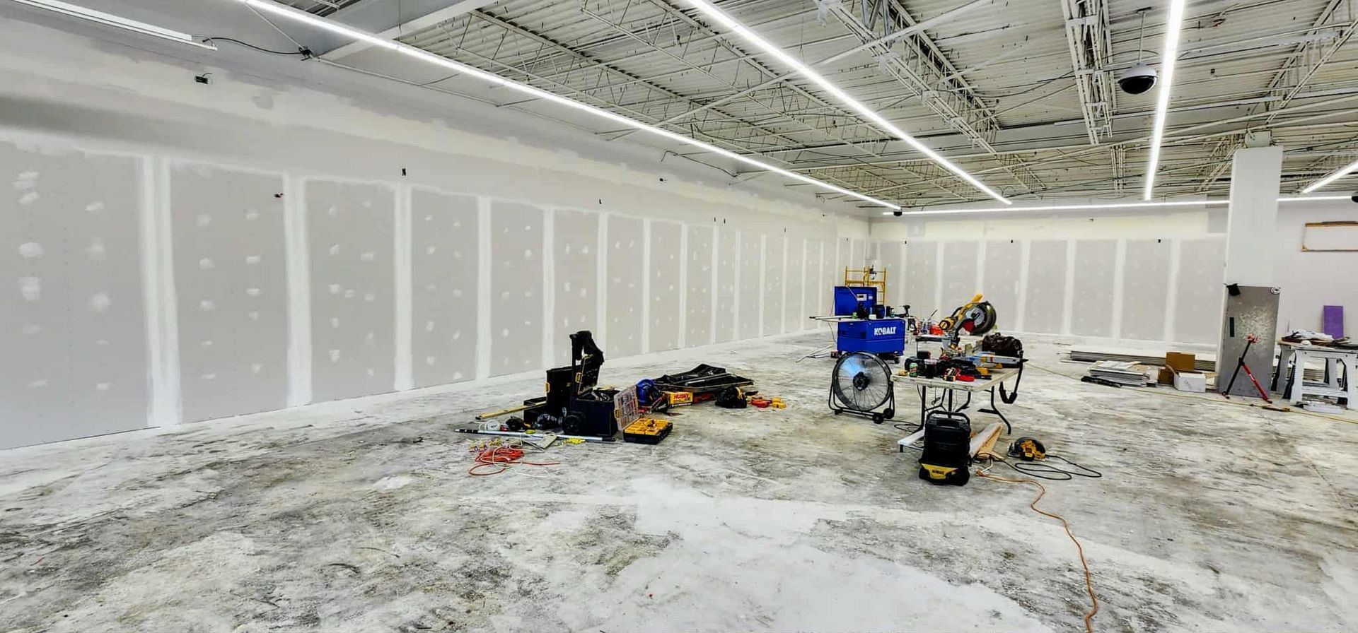 Interior of a commercial space under construction; drywall installation, tools, debris scattered on the floor.