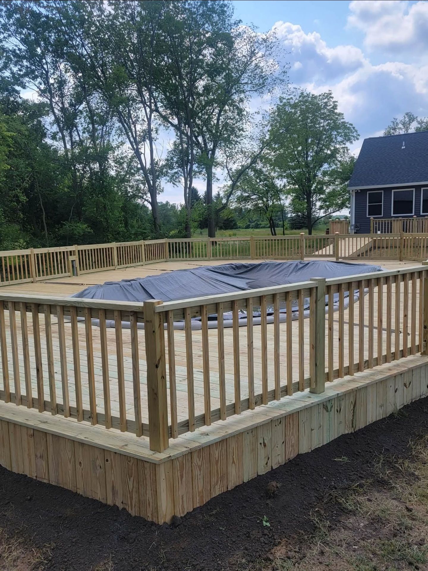 Wooden deck and fence surrounding a swimming pool with a black cover, in a grassy yard.