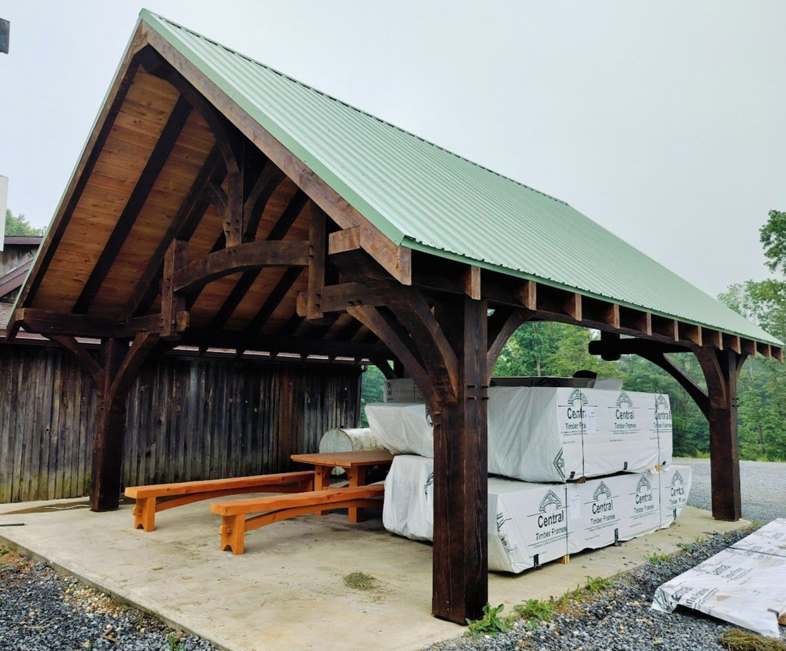 A timber-frame pavilion with a green metal roof, containing a wooden picnic table and stacked construction materials.