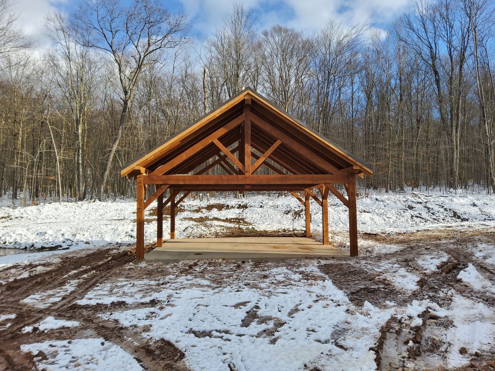 A wooden pavilion structure stands in a snowy, wooded clearing with muddy tire tracks in the foreground.