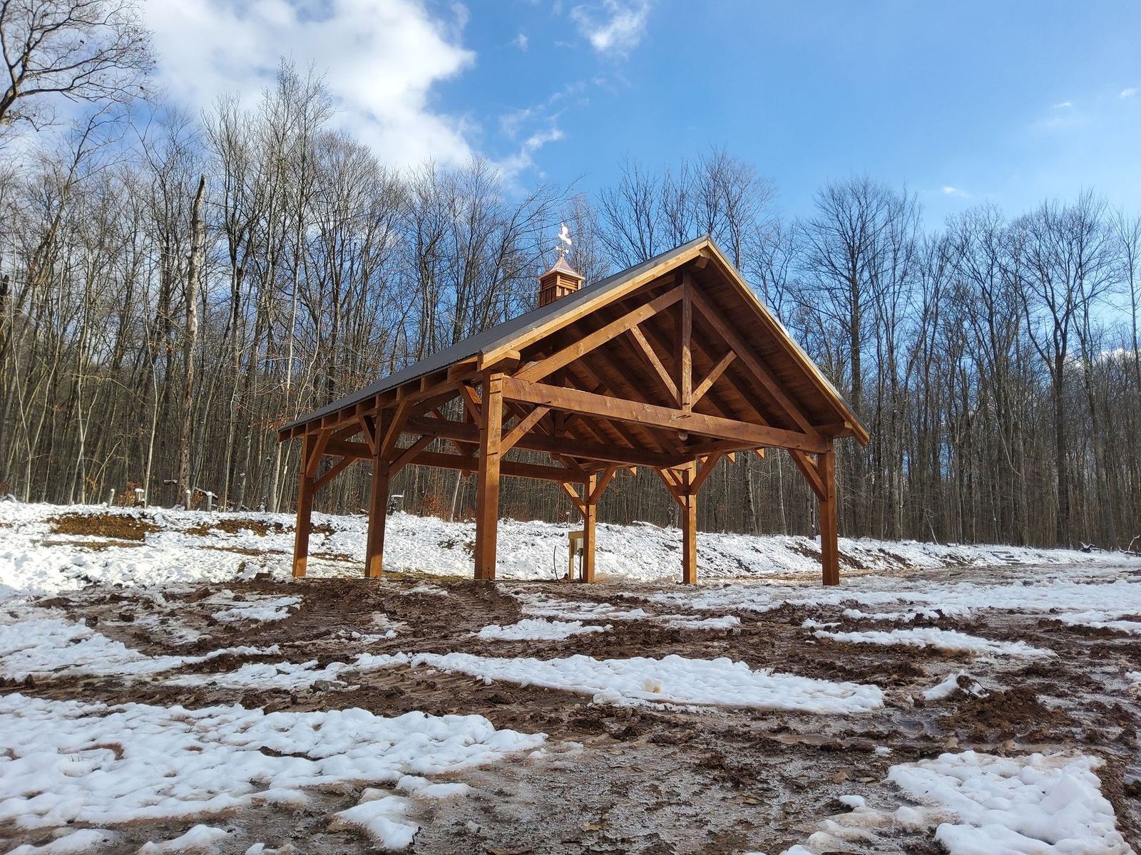 A rustic, open-air wooden pavilion sits on a snowy, mud-streaked lot in front of a dense, bare-branched forest.