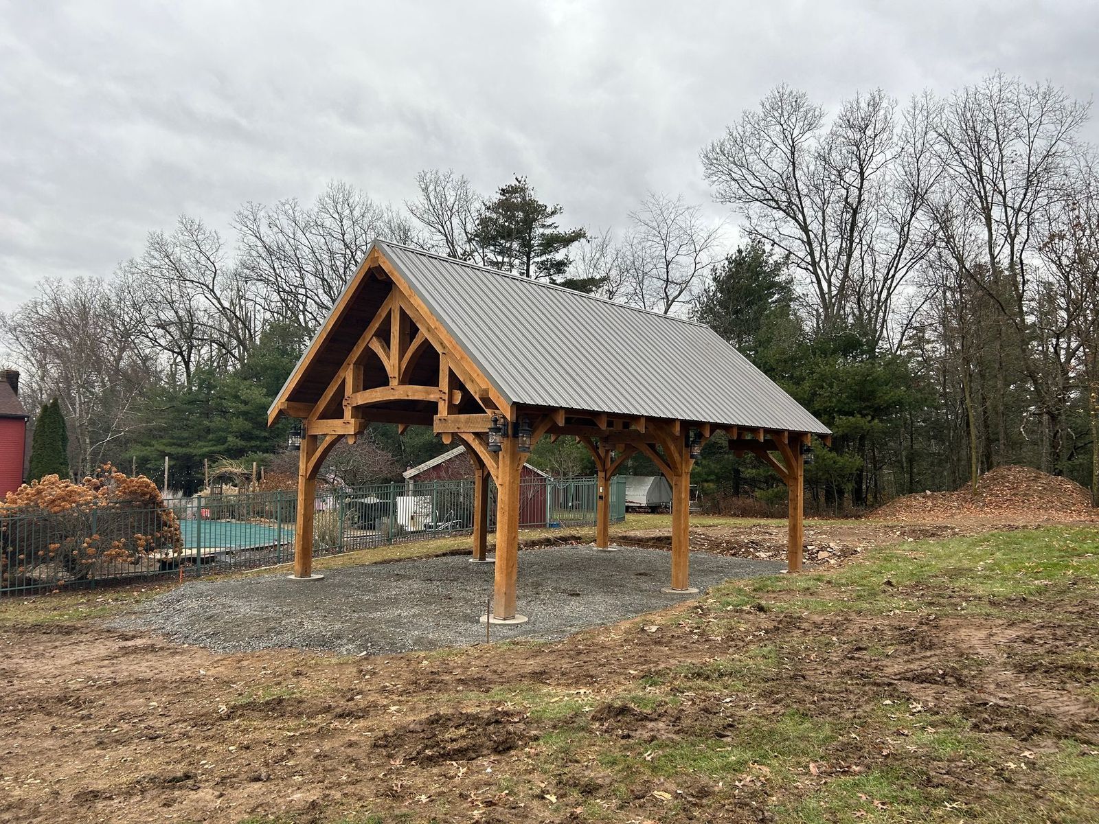 A timber-framed outdoor pavilion with a metal roof stands over a gravel pad in a rural, wooded setting.
