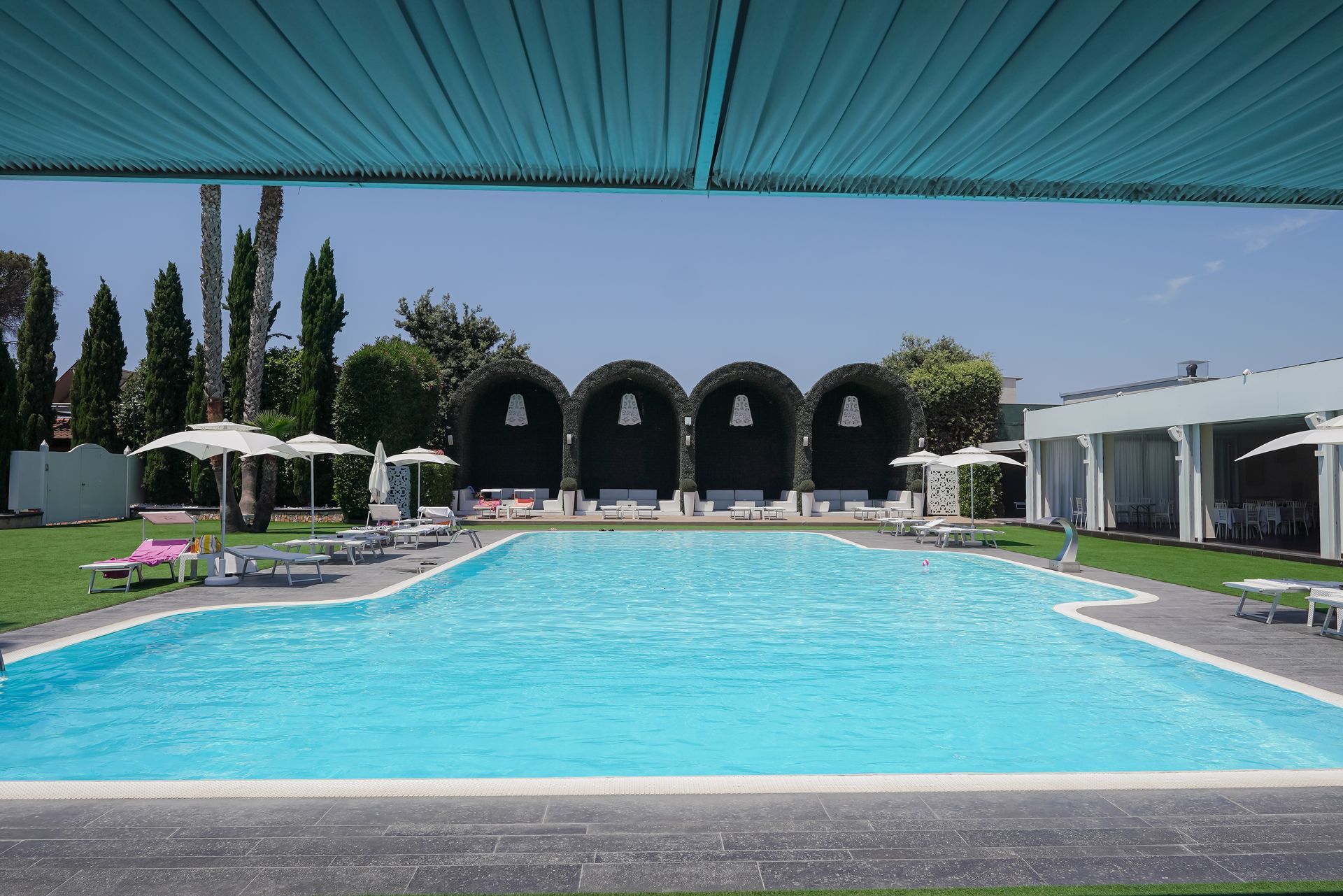 Piscina moderna del resort con acqua turchese, cabine e lettini bianchi sotto una tettoia blu.