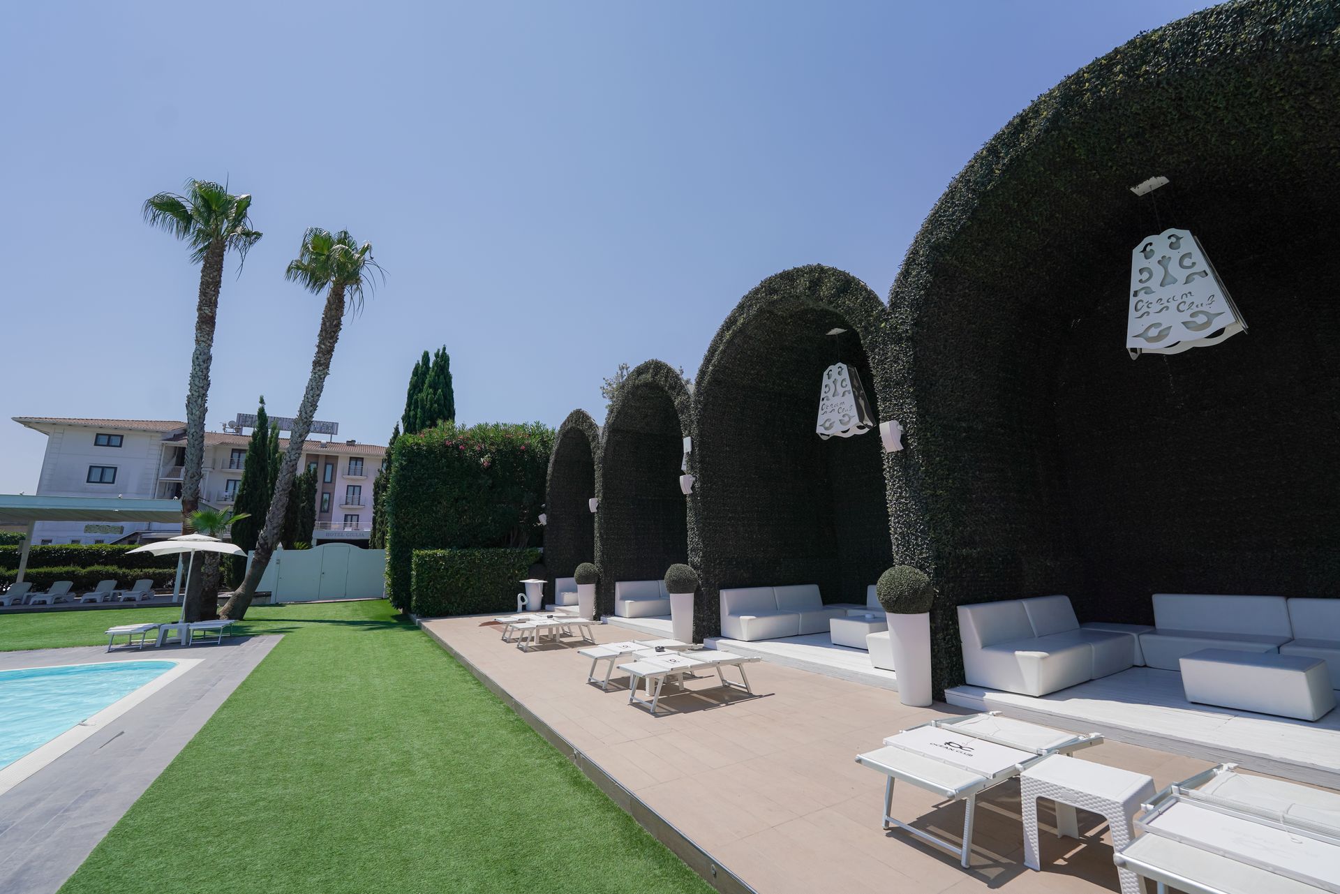 Salotto a bordo piscina con sedie bianche, prato verde, archi di arte topiaria e alte palme sotto un cielo azzurro.