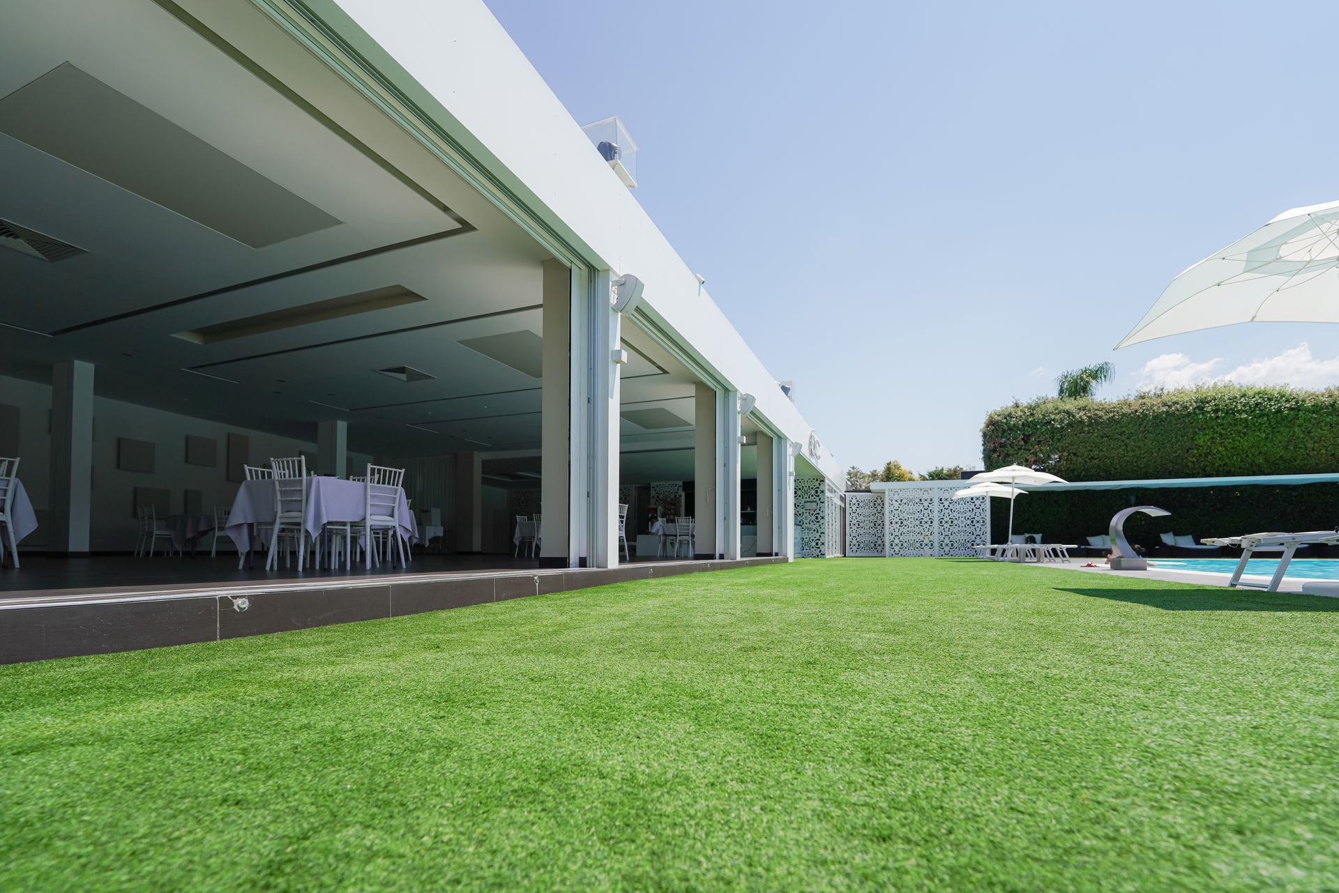 Patio di una casa moderna con porte scorrevoli aperte, sedie bianche, prato verde e una piscina sotto un cielo luminoso