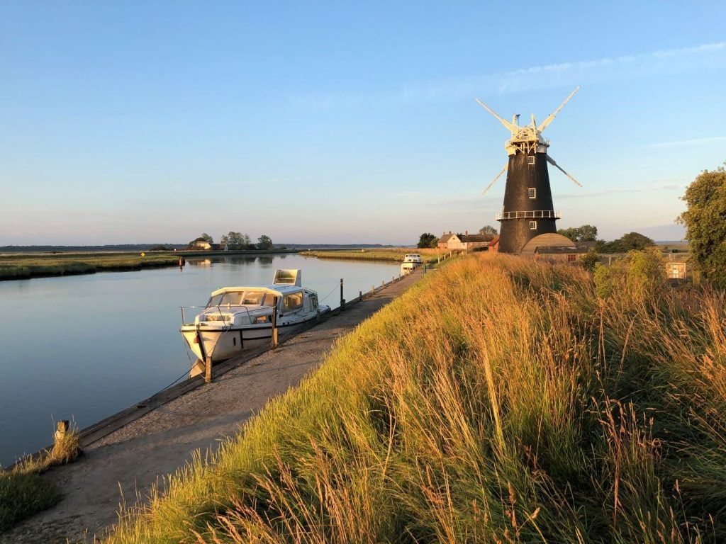 Boat docked on a riverbank next to a windmill, grass in foreground, blue sky.