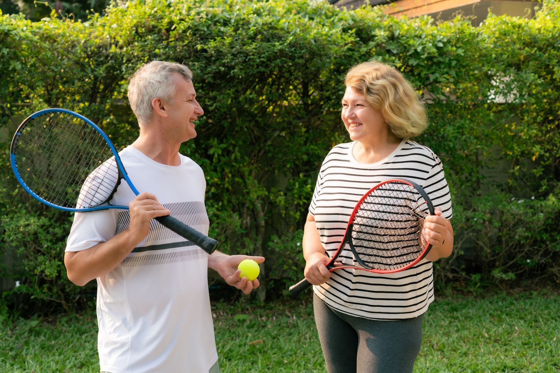 Man and woman with tennis rackets, smiling, outdoors. Man holds ball.