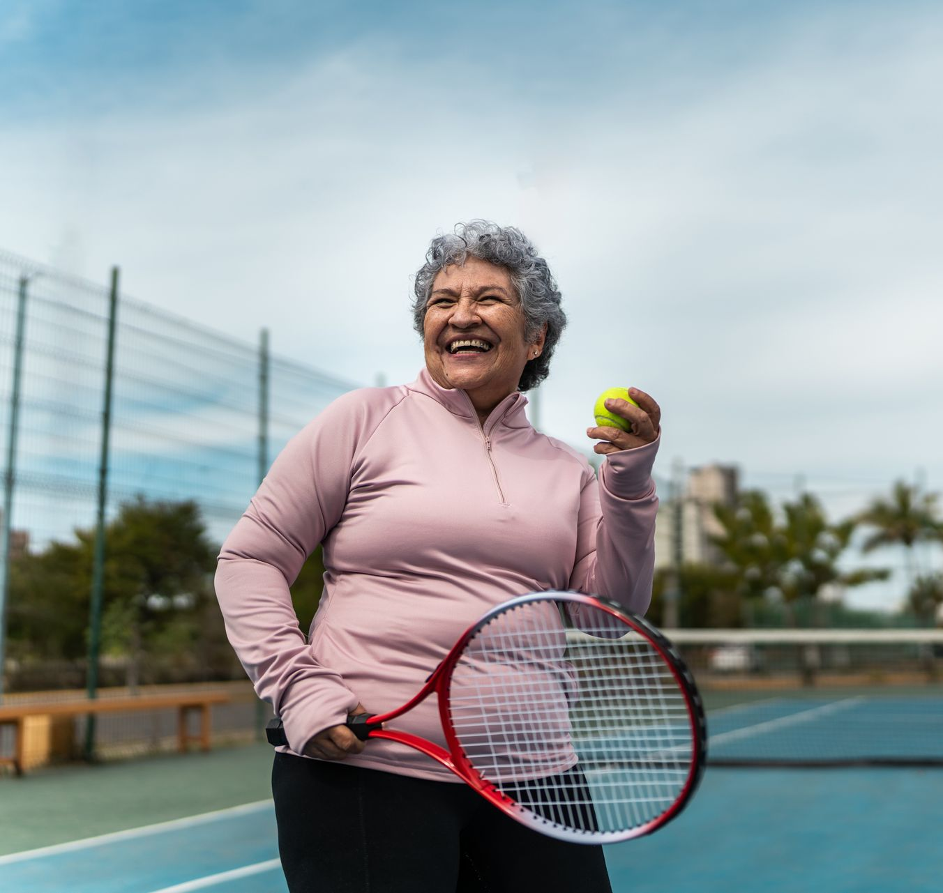An older woman with a tennis racket and ball smiles on a tennis court.