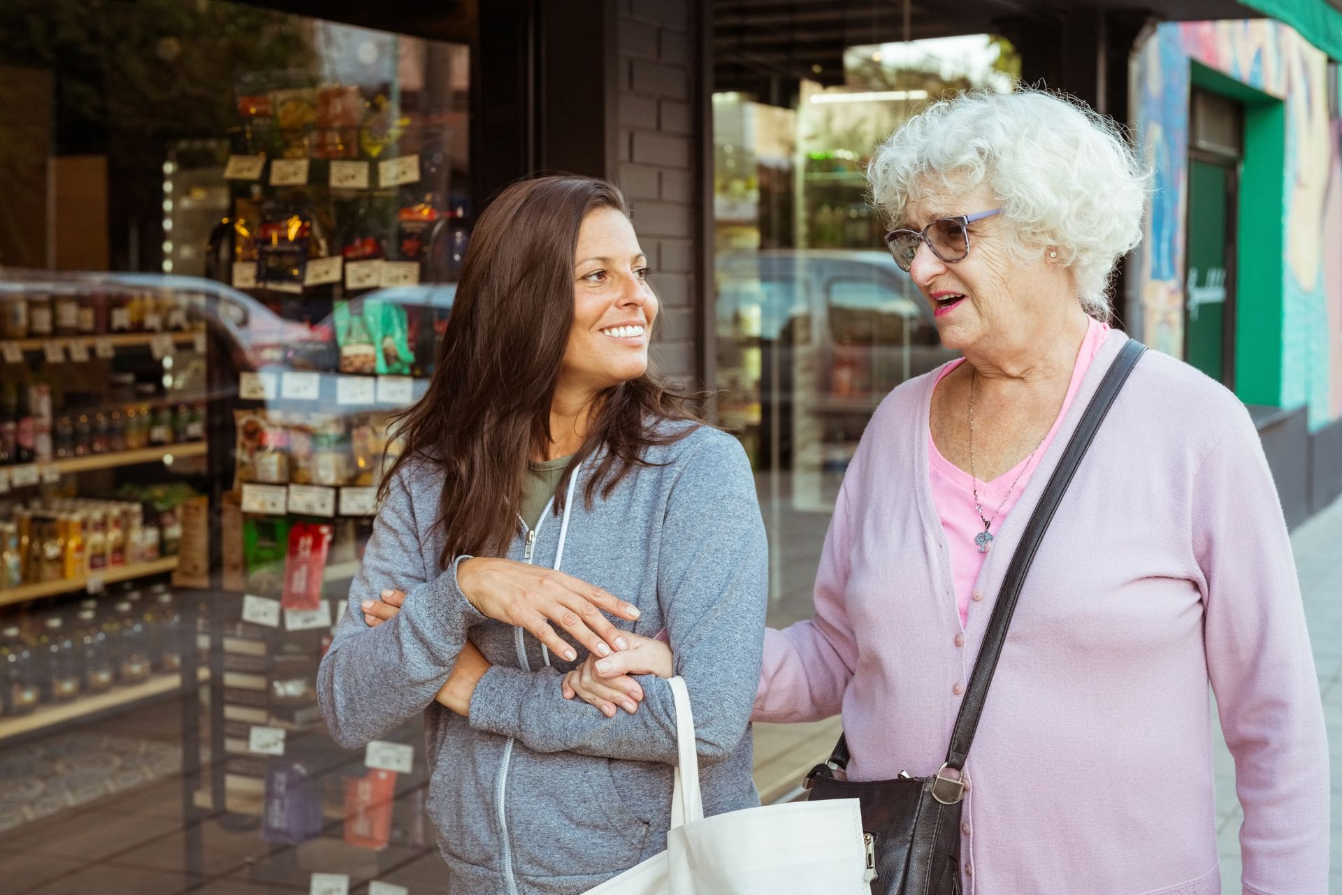 Woman smiles at another woman, who touches her arm on a sidewalk in front of a shop.