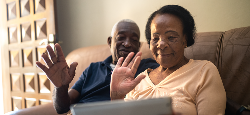 An older couple waving at a tablet, smiling, indoors on a couch.