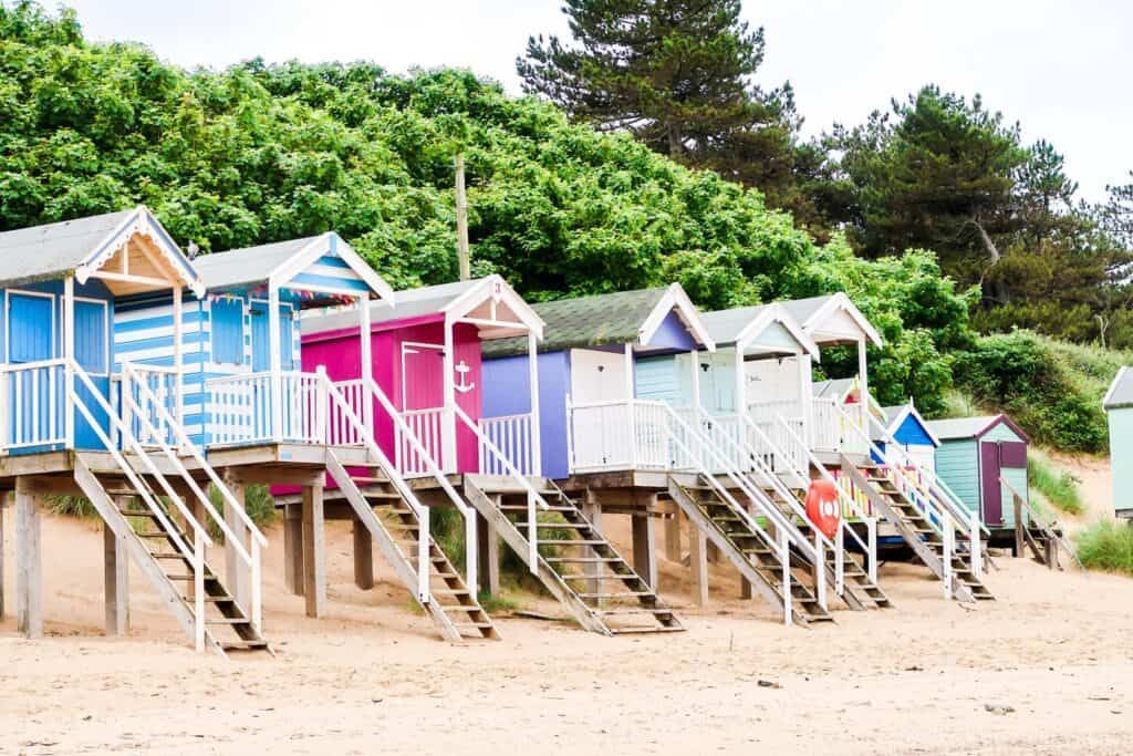 Colorful beach huts on stilts along a sandy beach, with green trees in the background.