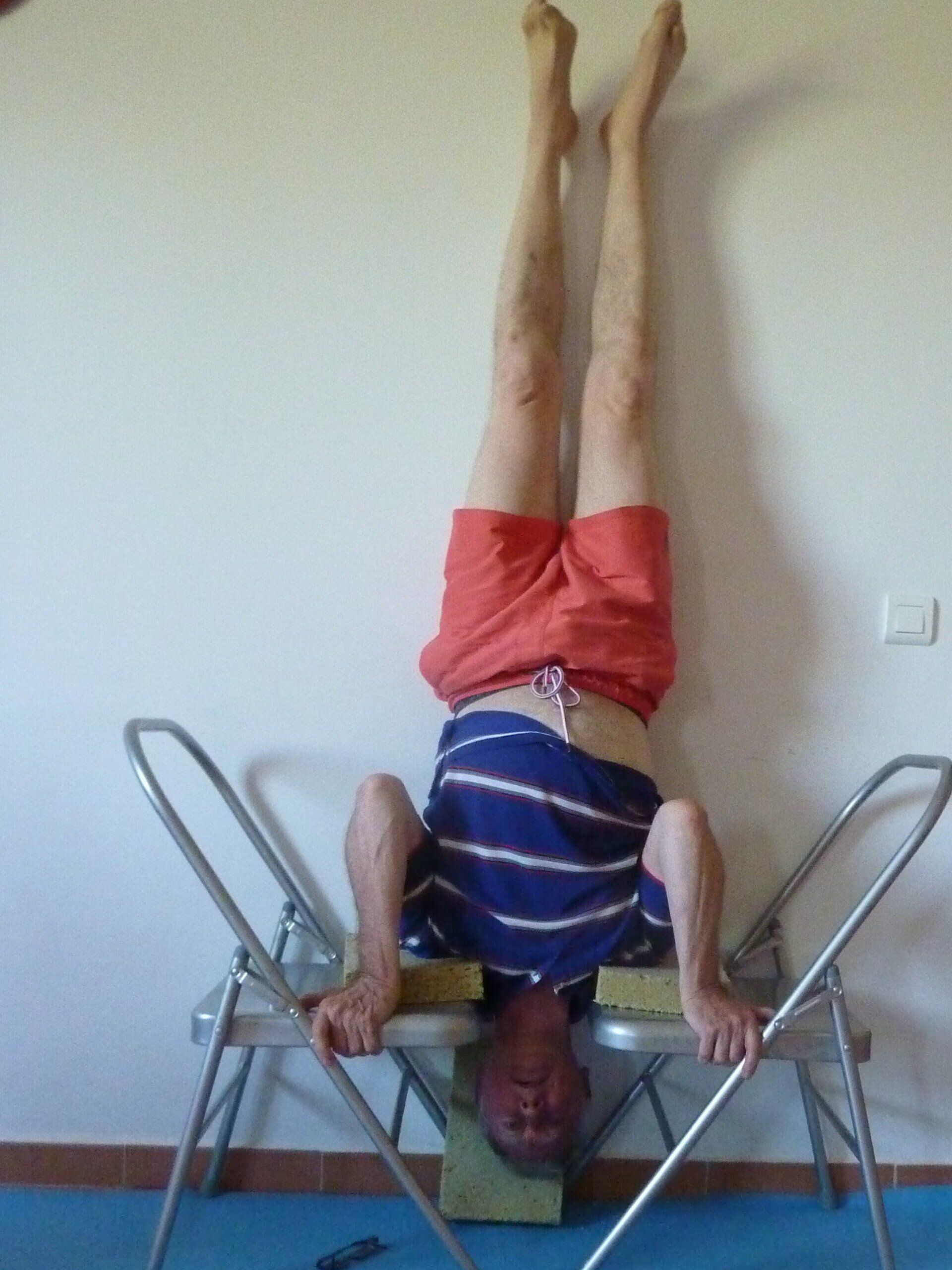 Man in headstand, using two chairs for support, against a white wall. He wears red shorts and a blue-striped shirt.