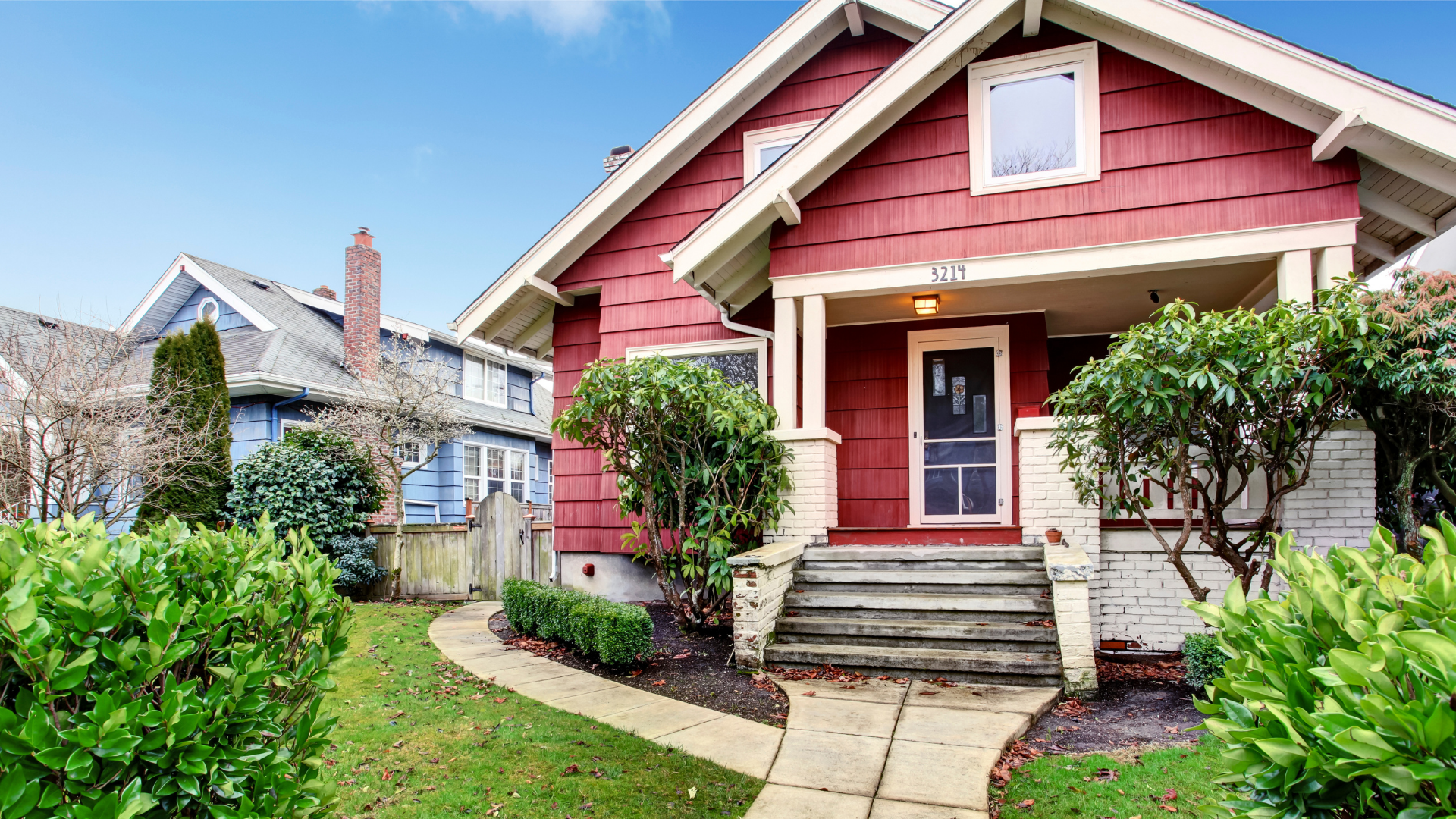 Red house with white trim, porch, and small front yard with bushes; blue house to the left.