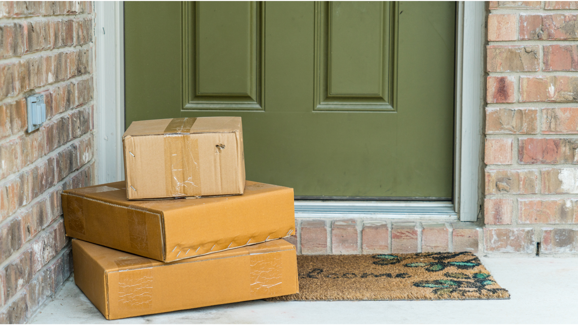 Three brown cardboard boxes stacked by a green door on a brick building's porch with a welcome mat.