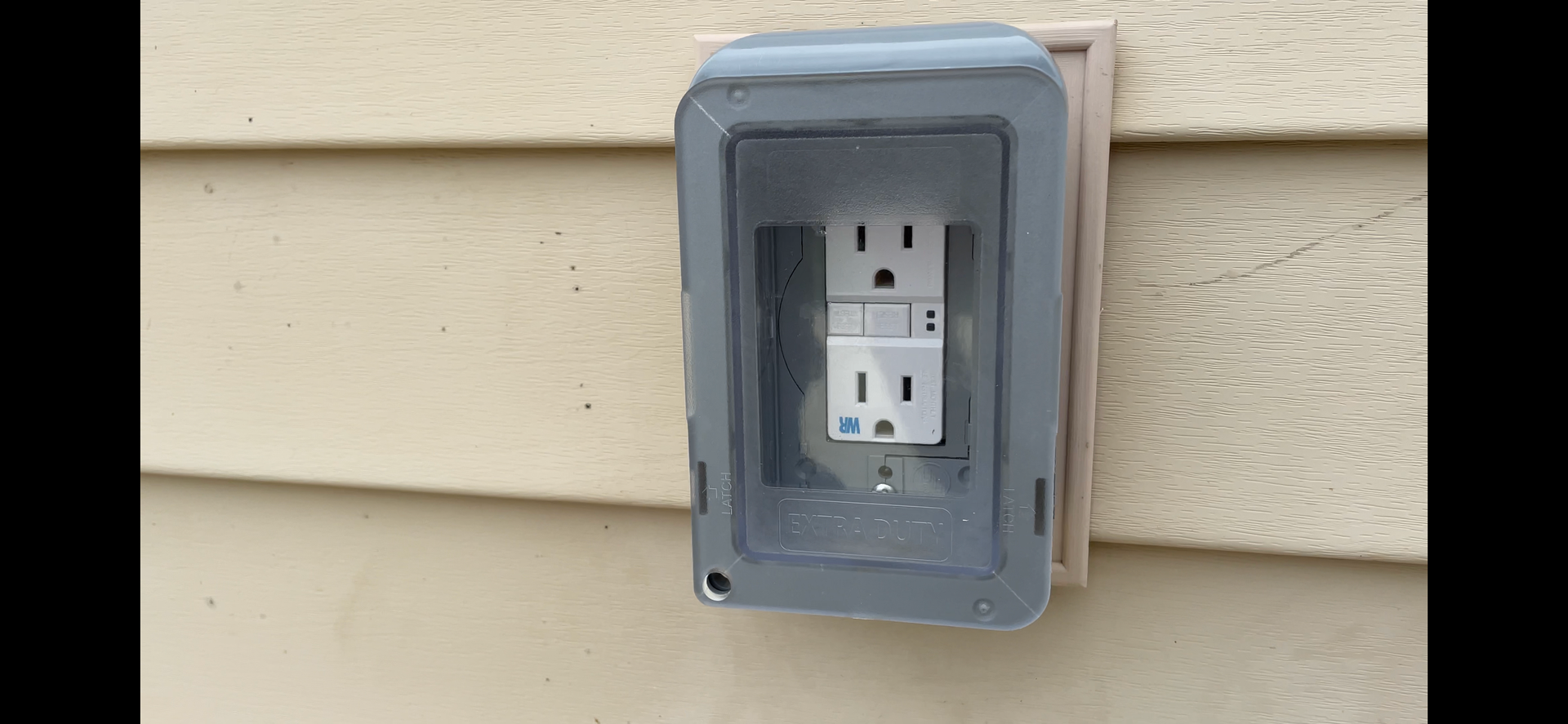 Outdoor electrical outlet inside a gray weatherproof cover, mounted on beige siding.