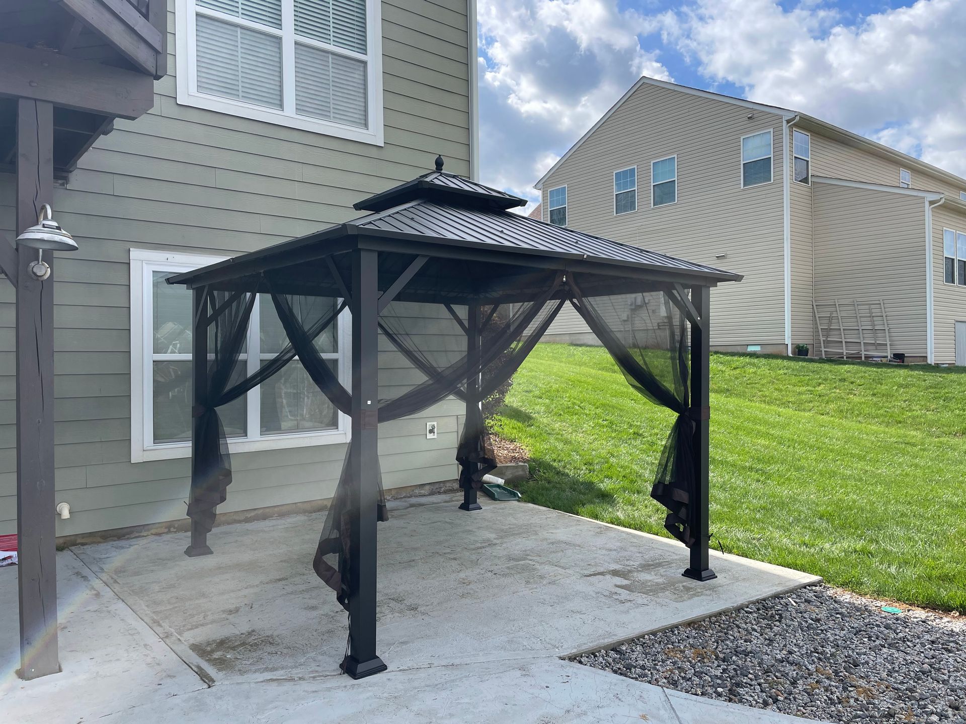 Black gazebo with curtains on a concrete patio, with a grassy backyard and house.