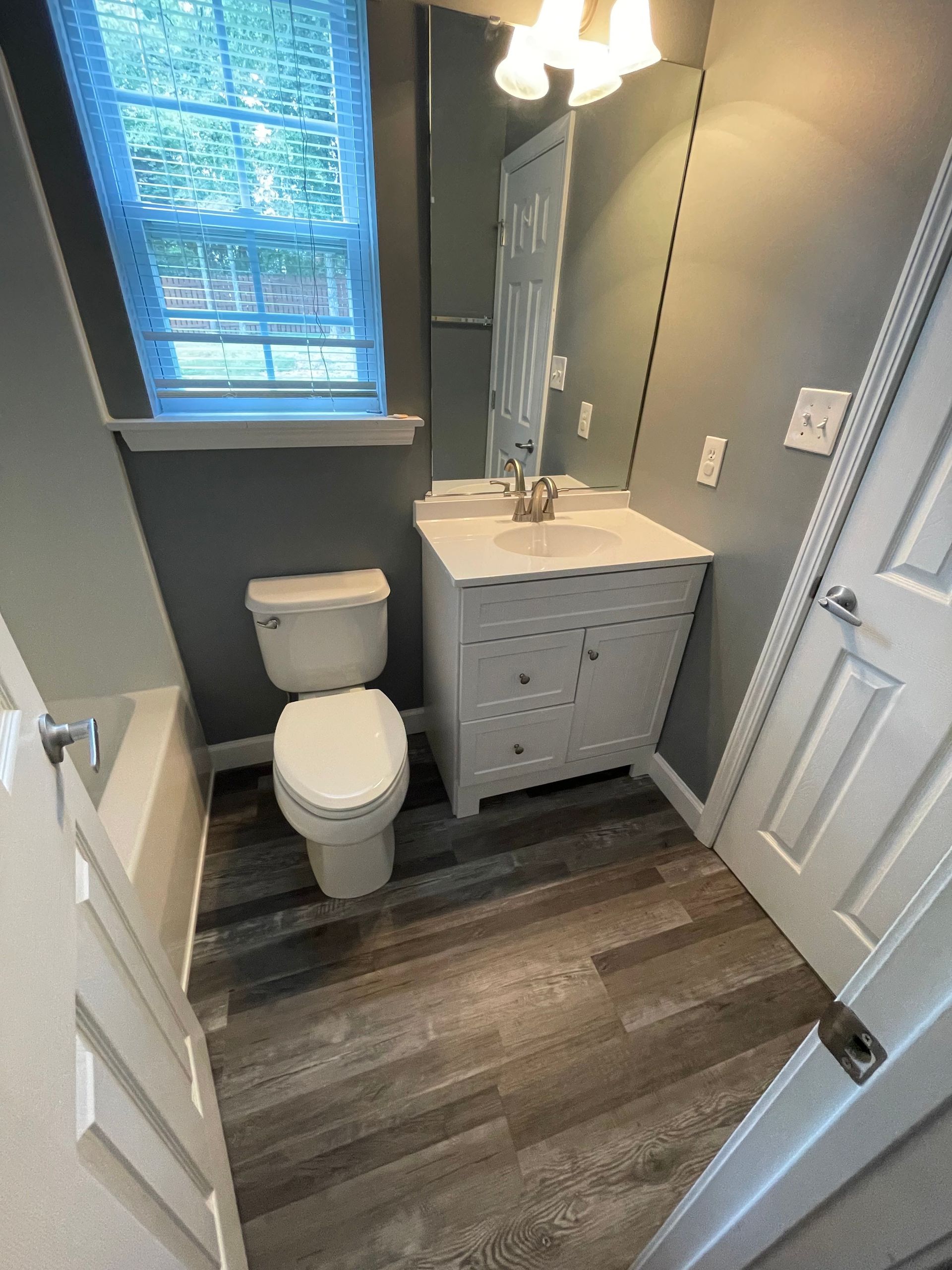 Small bathroom with a toilet, sink, and window. Grey walls, dark wood-look flooring, white cabinet.