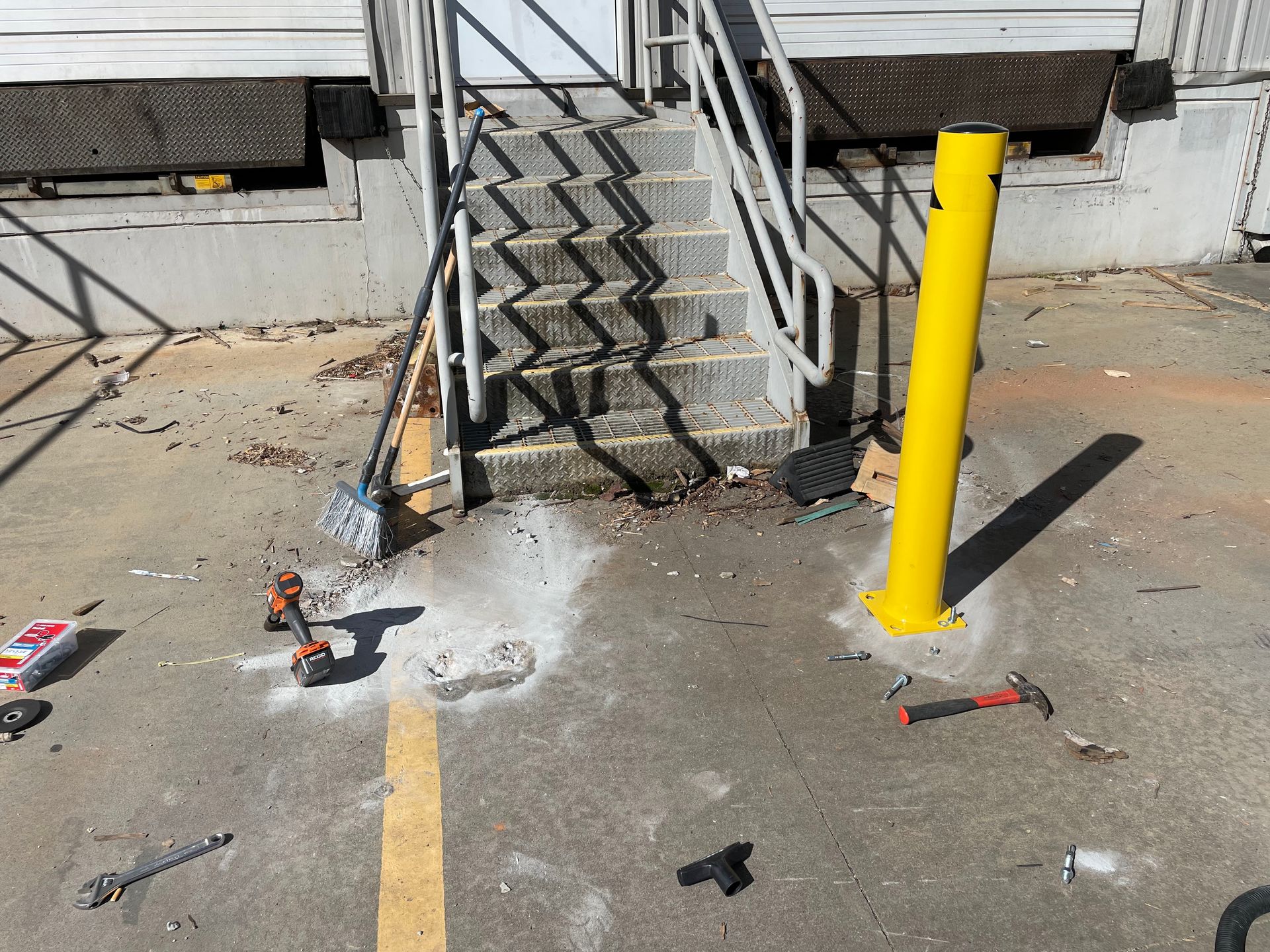 A yellow bollard by stairs leading to a loading dock; tools and debris litter the ground.