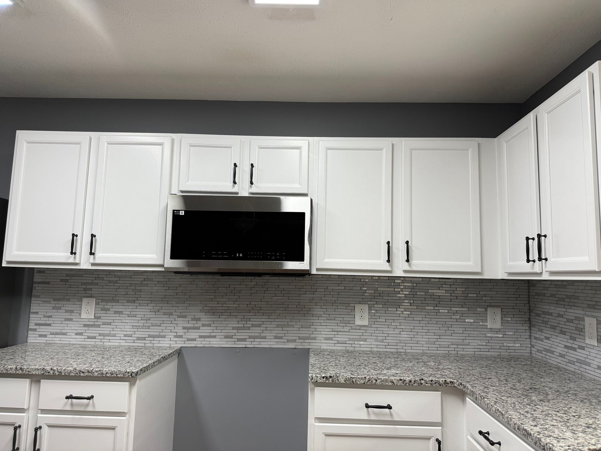 White kitchen cabinets, microwave, and countertop with black hardware, gray walls, and granite backsplash.