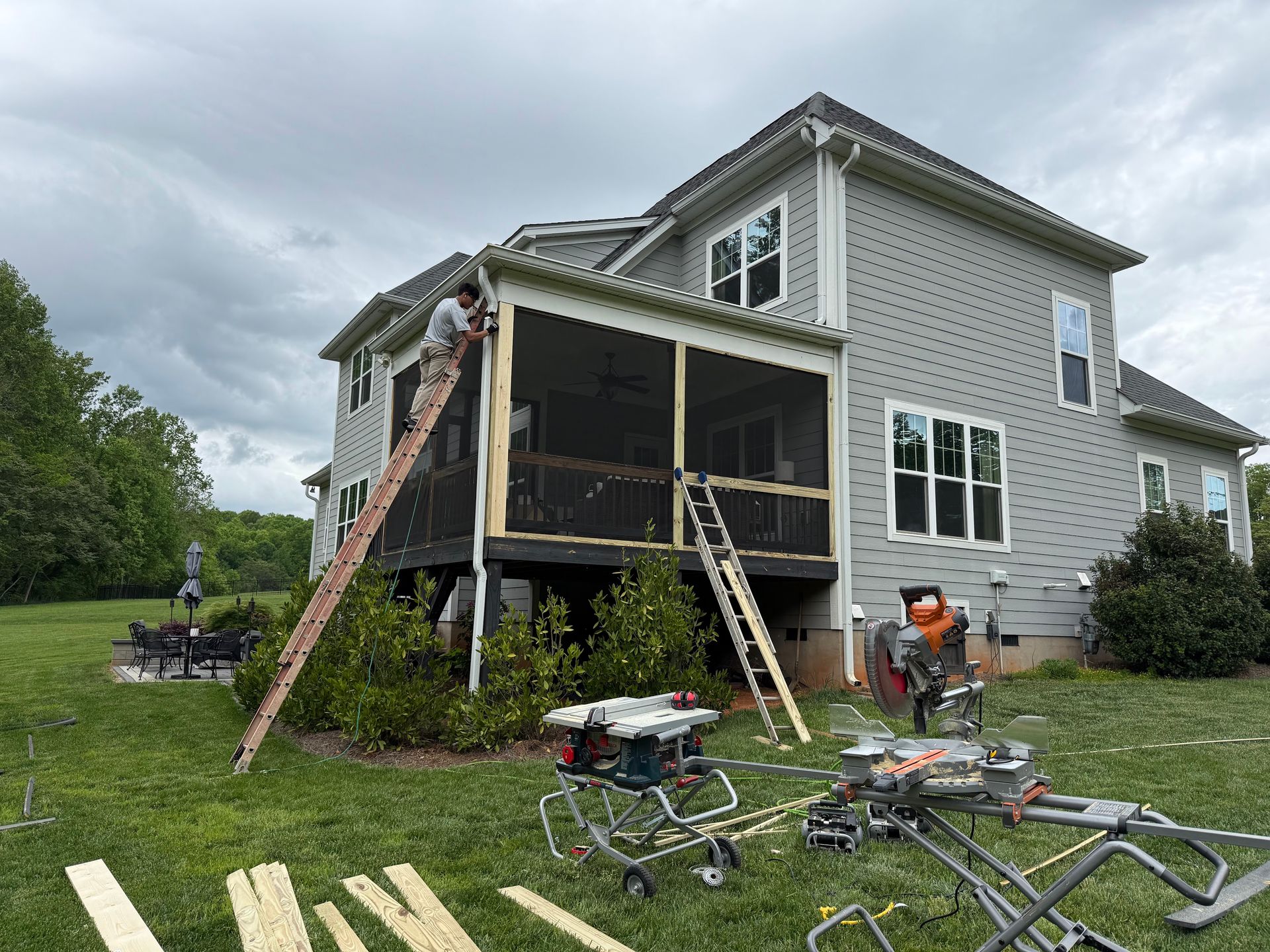 Man on ladder building a screened porch on a gray house; tools and materials on the grass.