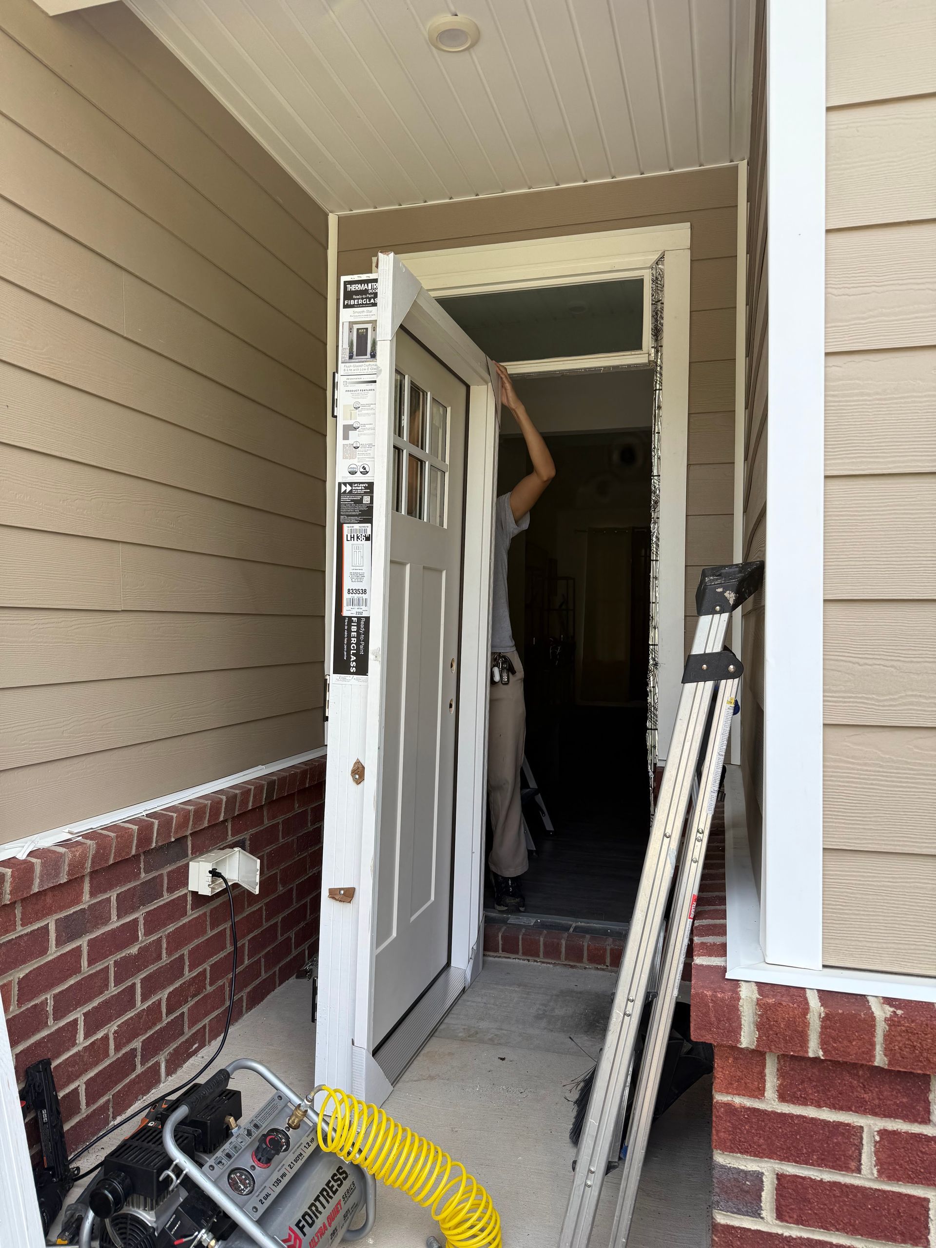 Person installing a white door in a tan building with a brick base. A ladder leans against the frame.