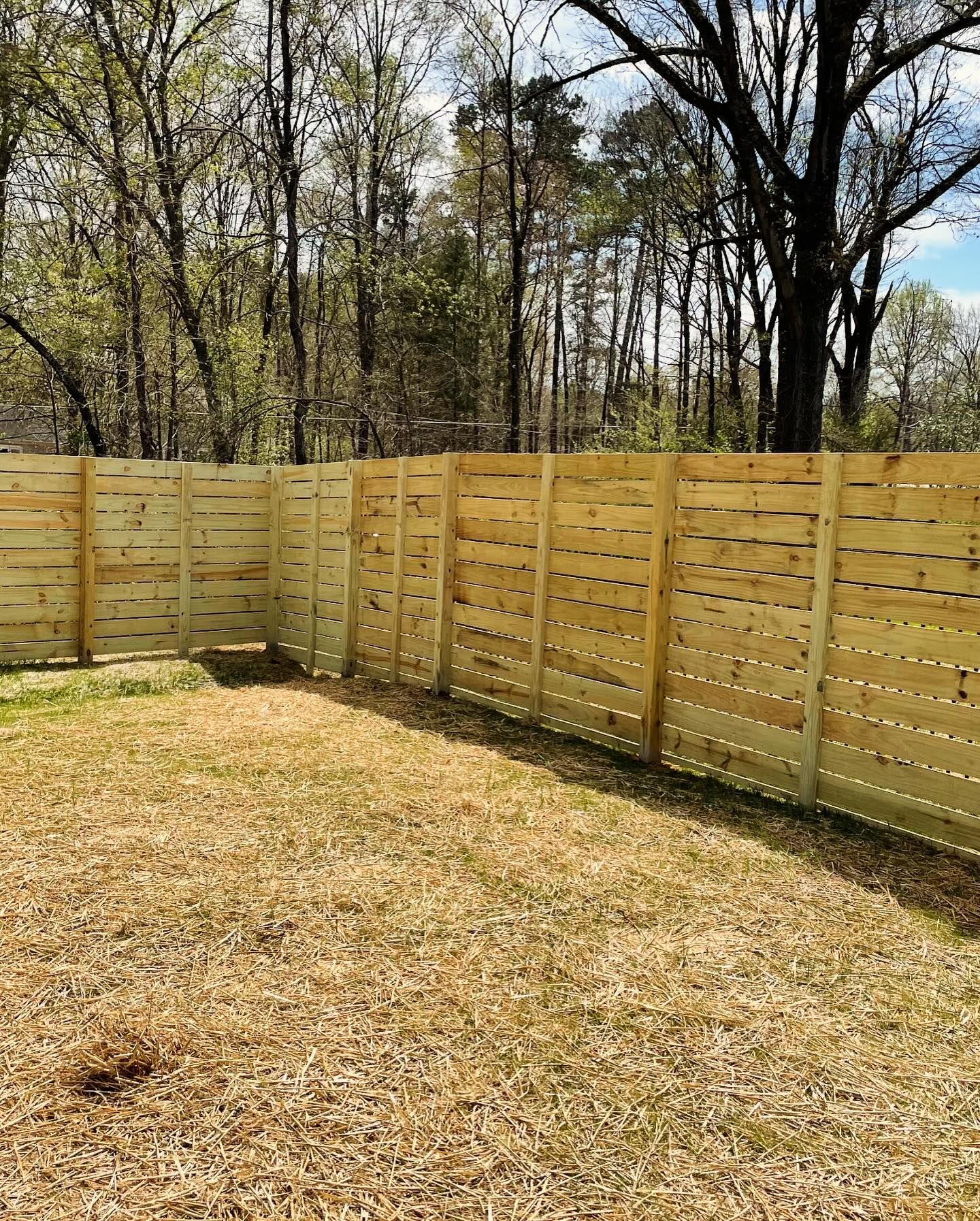 Wooden horizontal slat fence in a yard with brown grass and trees in the background.