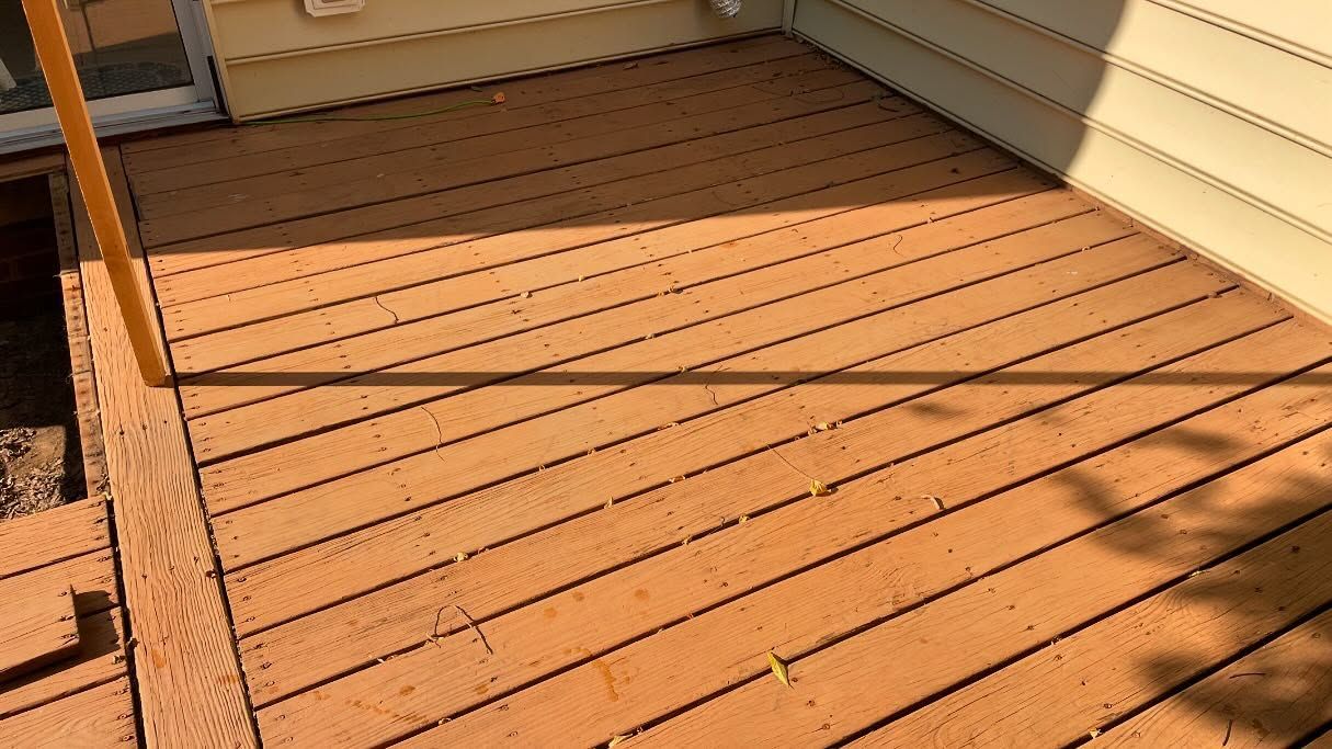 Wooden deck with orange-brown planks. Sunlight casts shadows. Yellow pole on left. House siding on right.