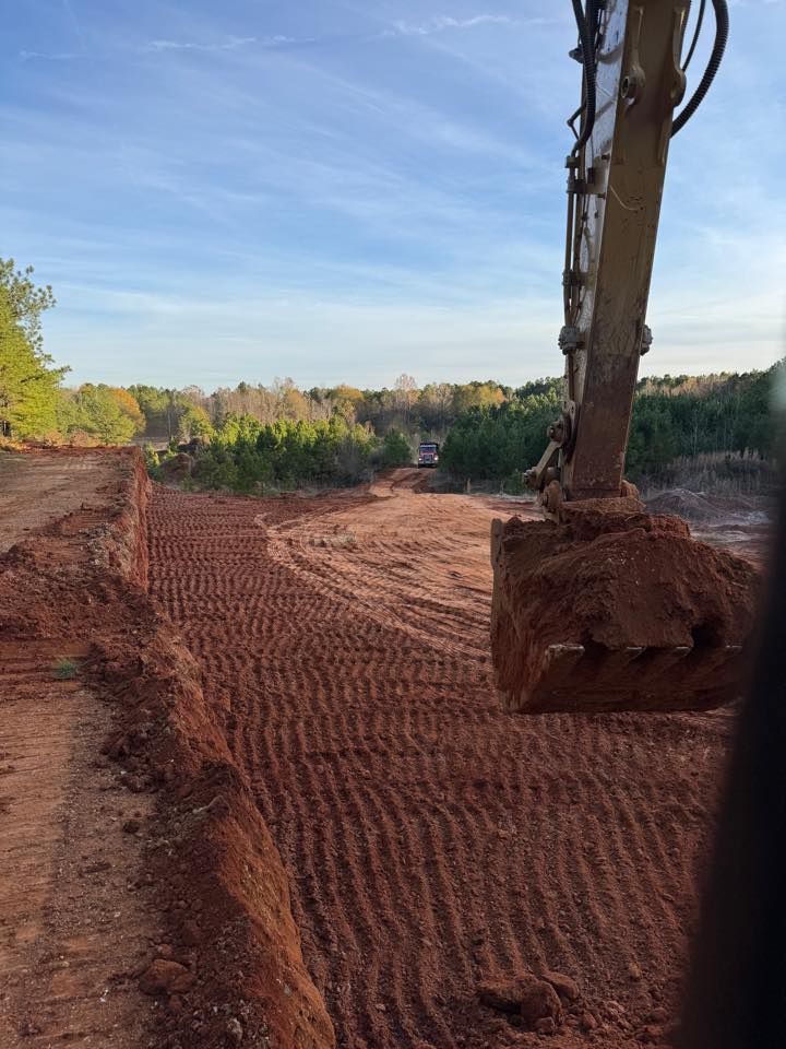 Excavator digging a trench in red dirt, trees in the background, under a blue sky.