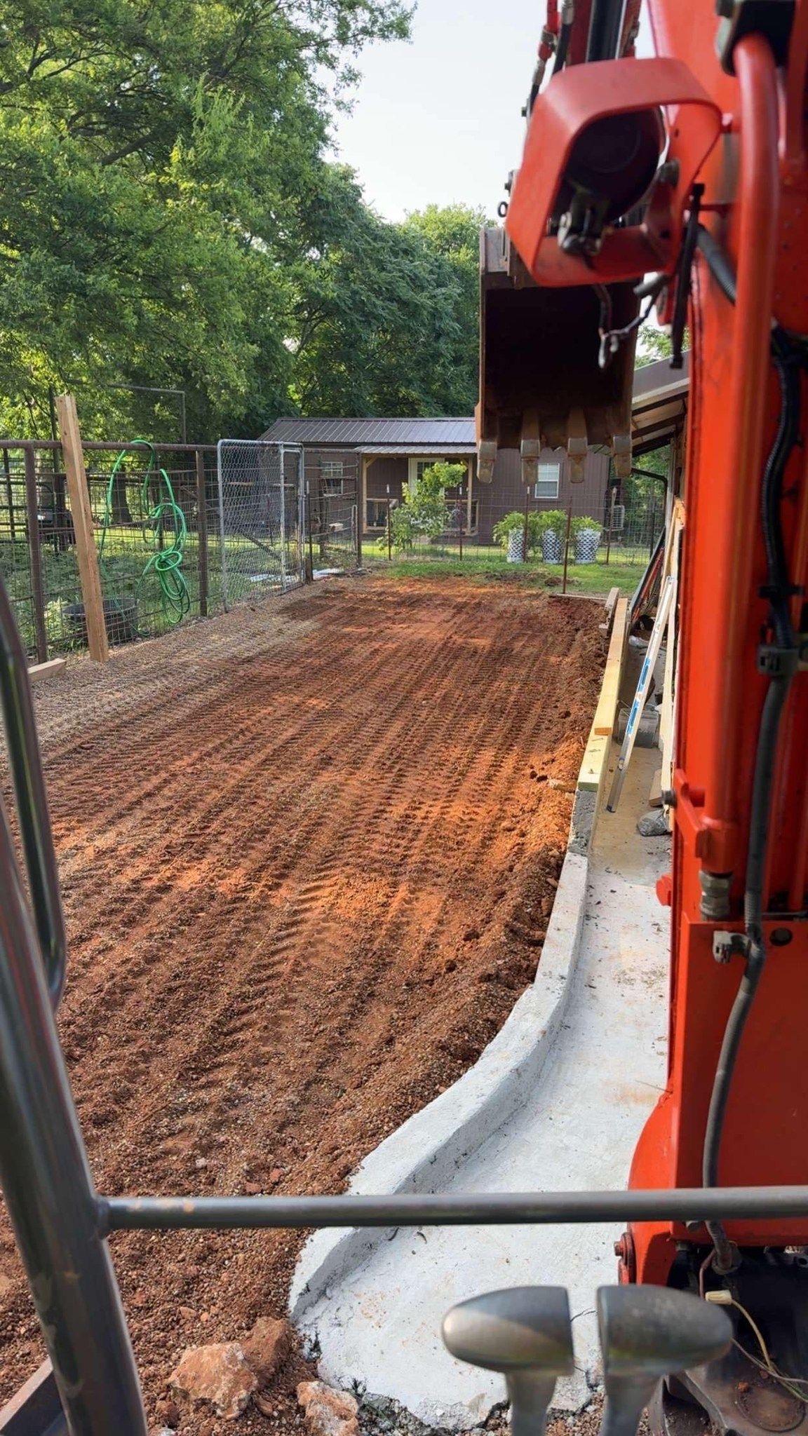 Excavator with bucket over dirt path next to concrete curb. Building and trees in the background.