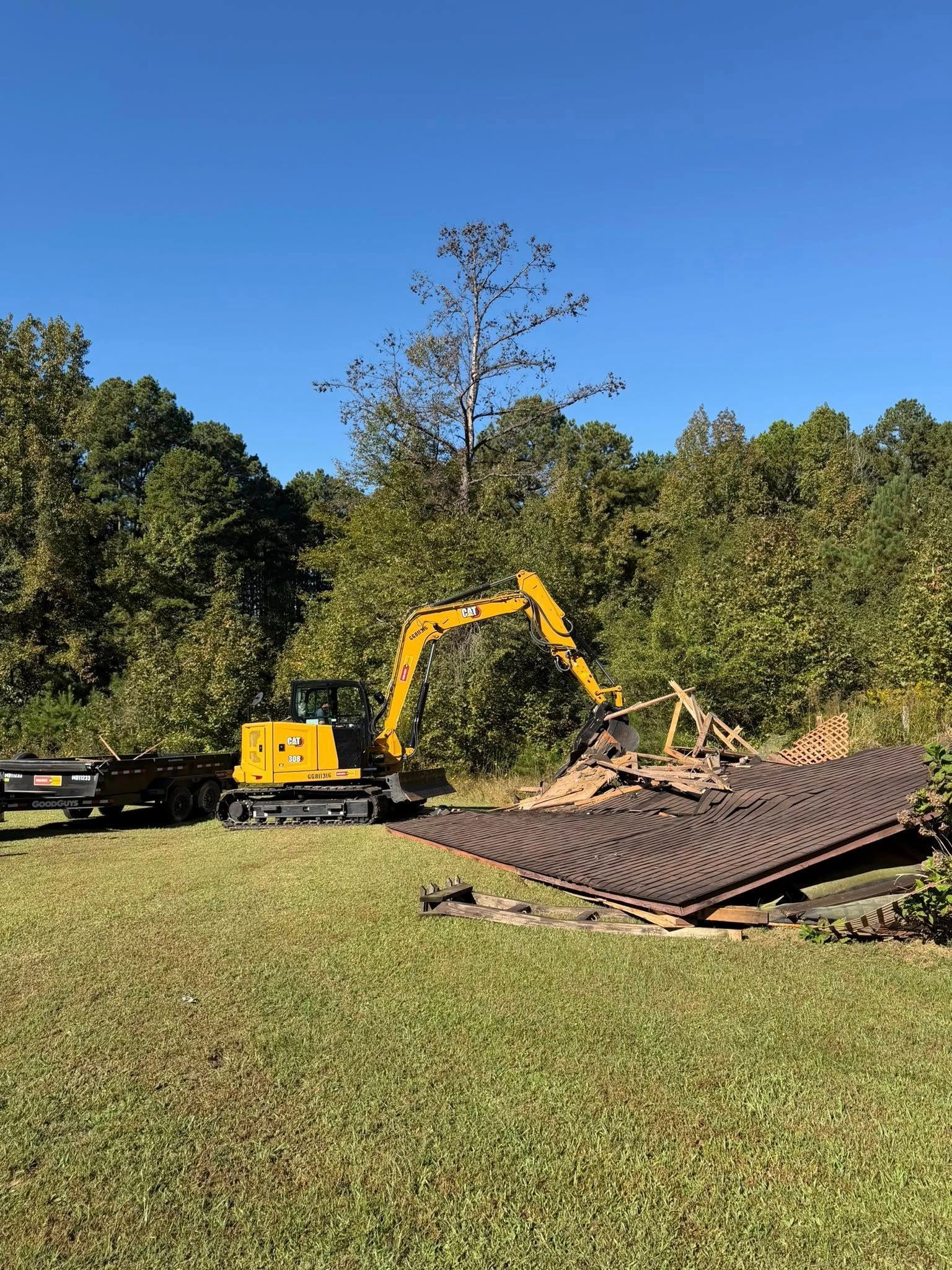 Yellow excavator demolishes a roof on a grassy area surrounded by trees under a blue sky.