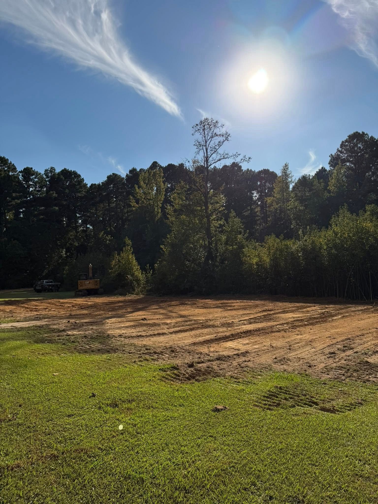 Cleared dirt patch with green grass foreground, trees, and bright sun in a blue sky.