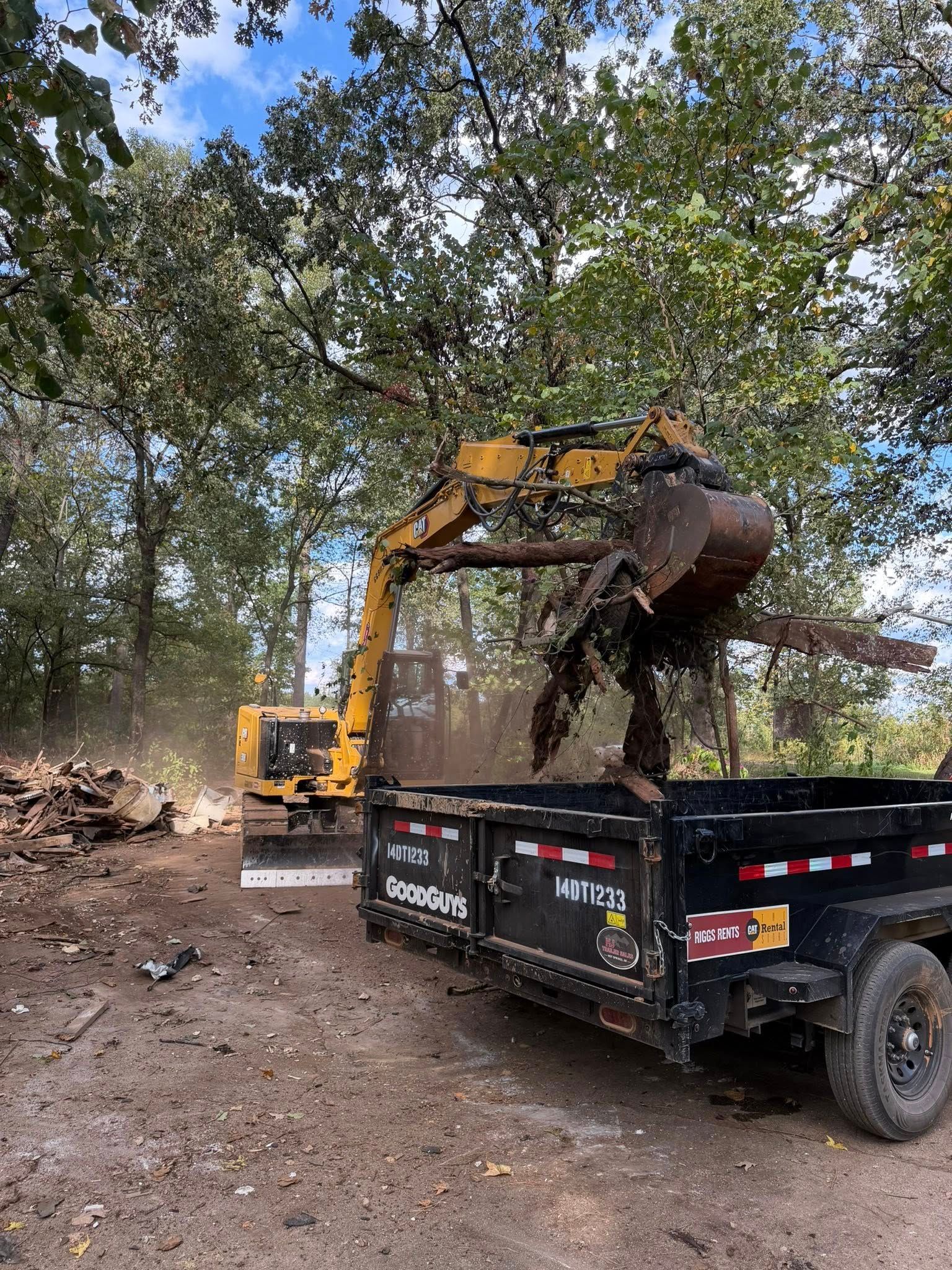 Yellow excavator loading debris into a black trailer in a wooded area under a blue sky.