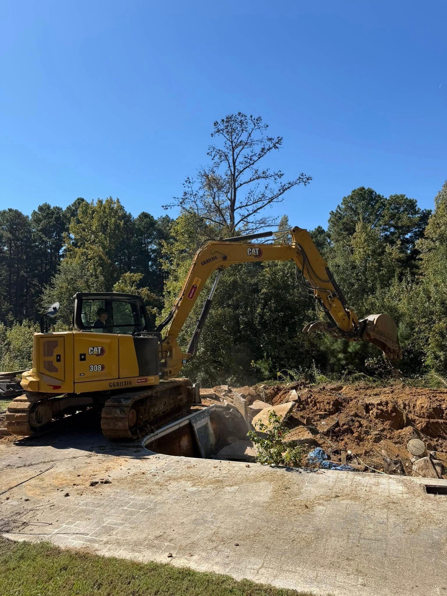 Yellow excavator digging in a dirt pile, set against trees and a blue sky.