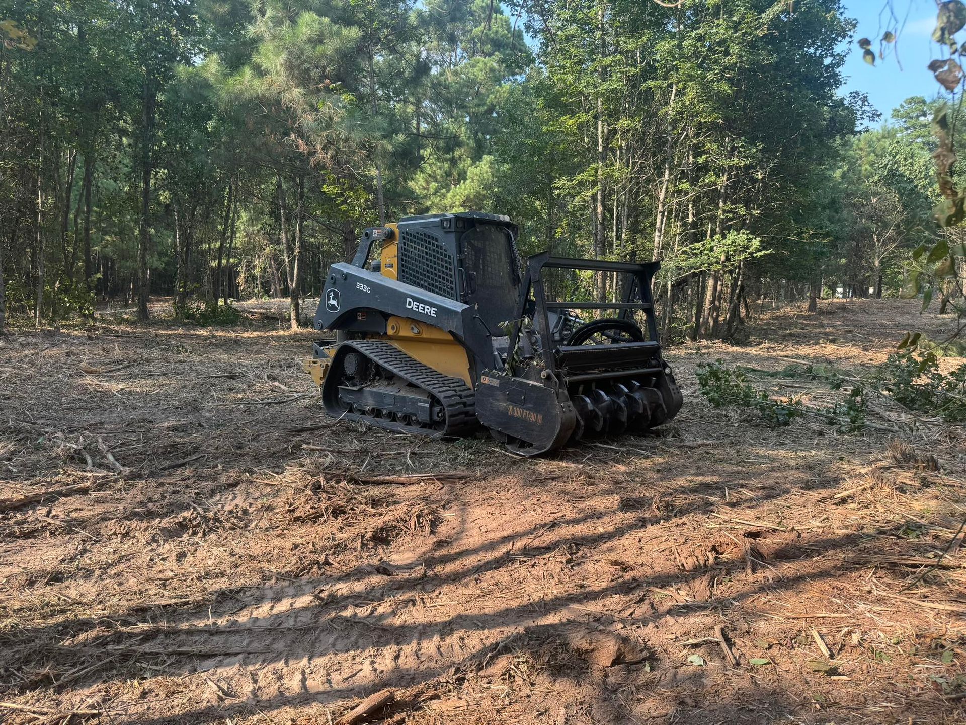 A yellow and black skid-steer loader with a mulcher attachment clearing land in a forest setting.