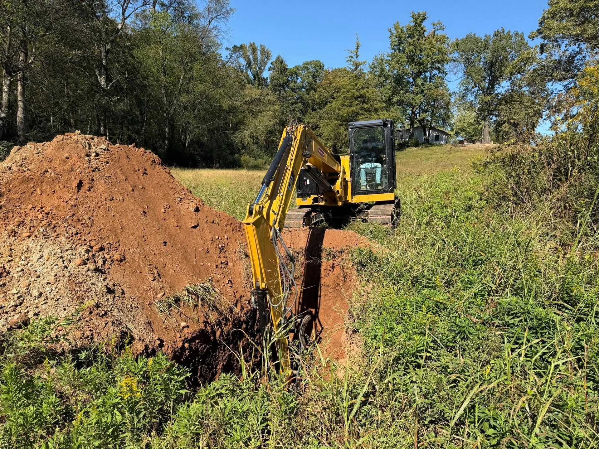 Yellow excavator digging a trench in grassy field next to a dirt pile. Trees and sky in background.