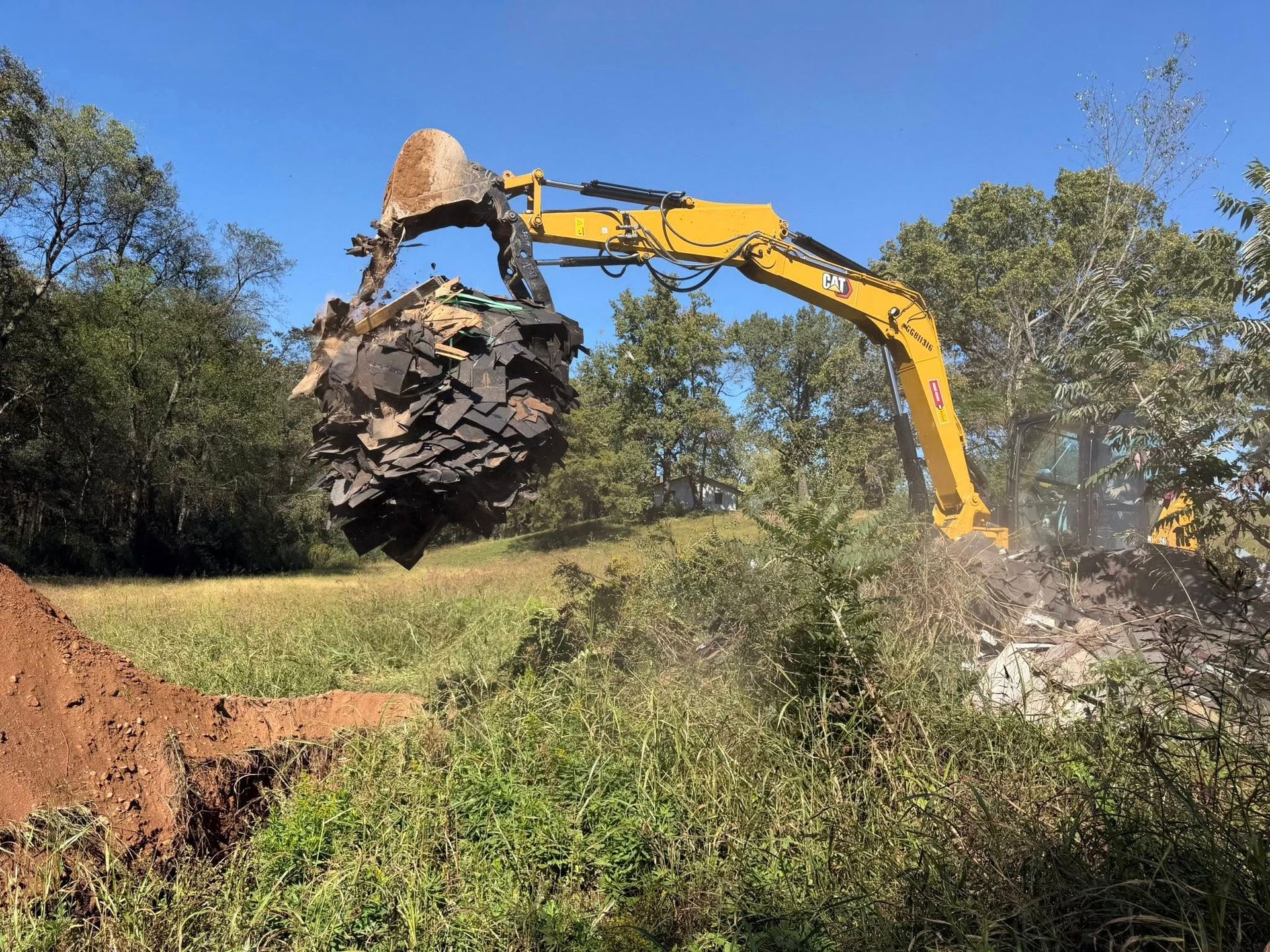 Yellow excavator lifting a large, dark pile of debris from a trench in a grassy field under a blue sky.