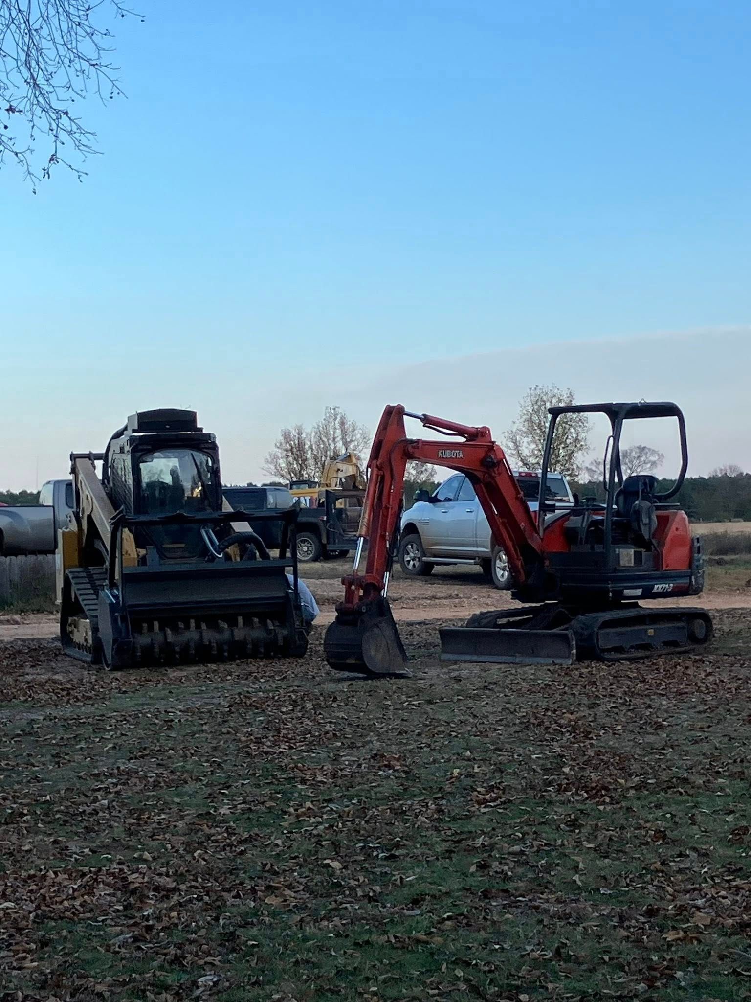Two construction vehicles on a grassy field. A black skid steer loader and a small orange excavator. Cloudy sky.