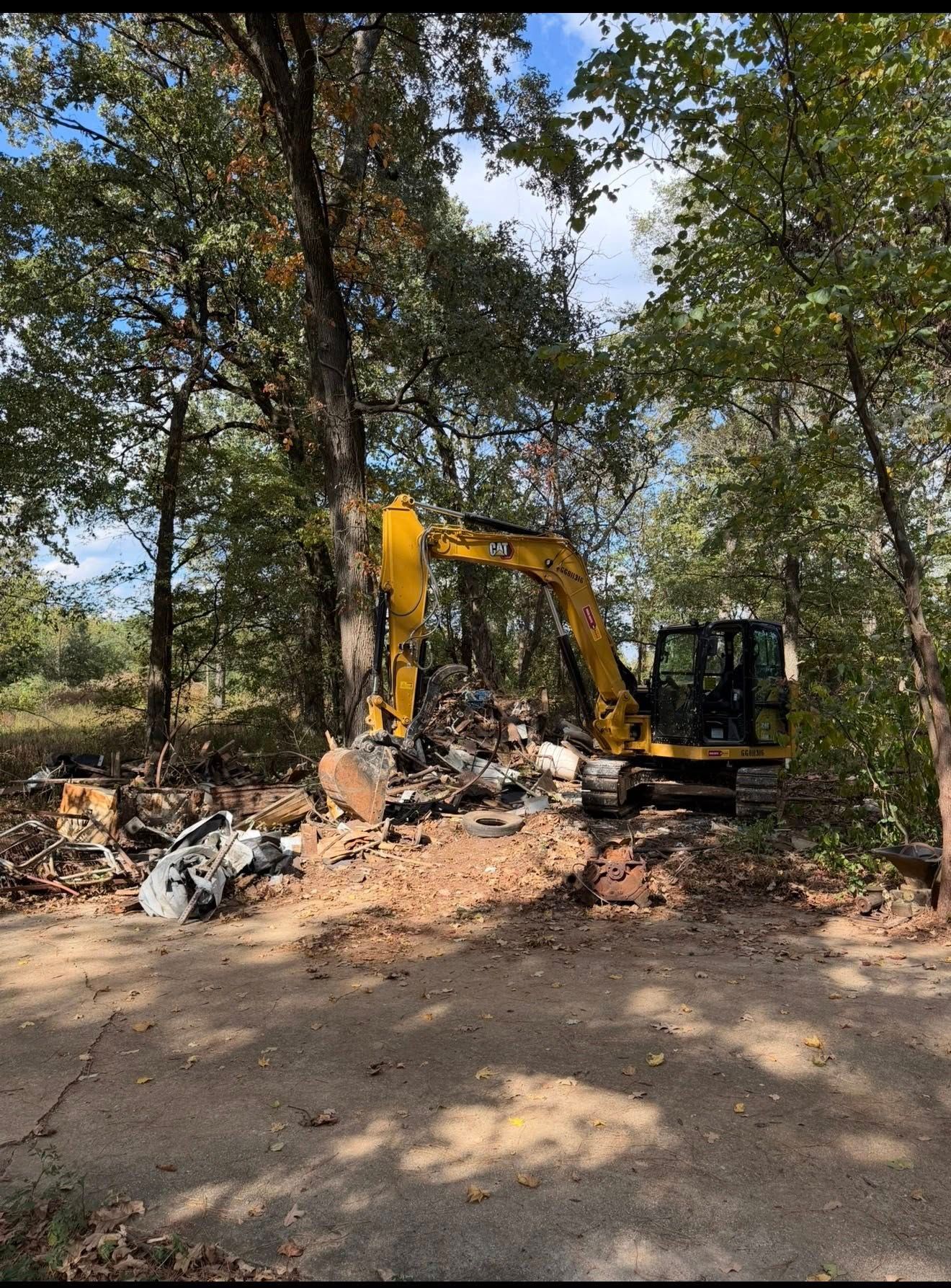 Yellow excavator clearing debris and rocks in a wooded area on a sunny day.