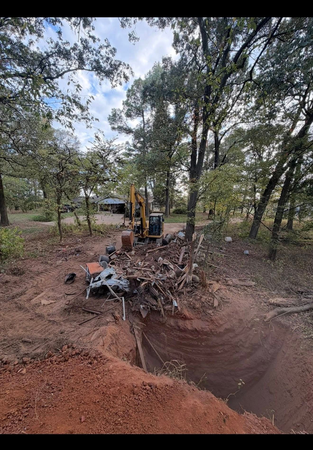 An excavator clearing debris in a wooded area with a large trench.