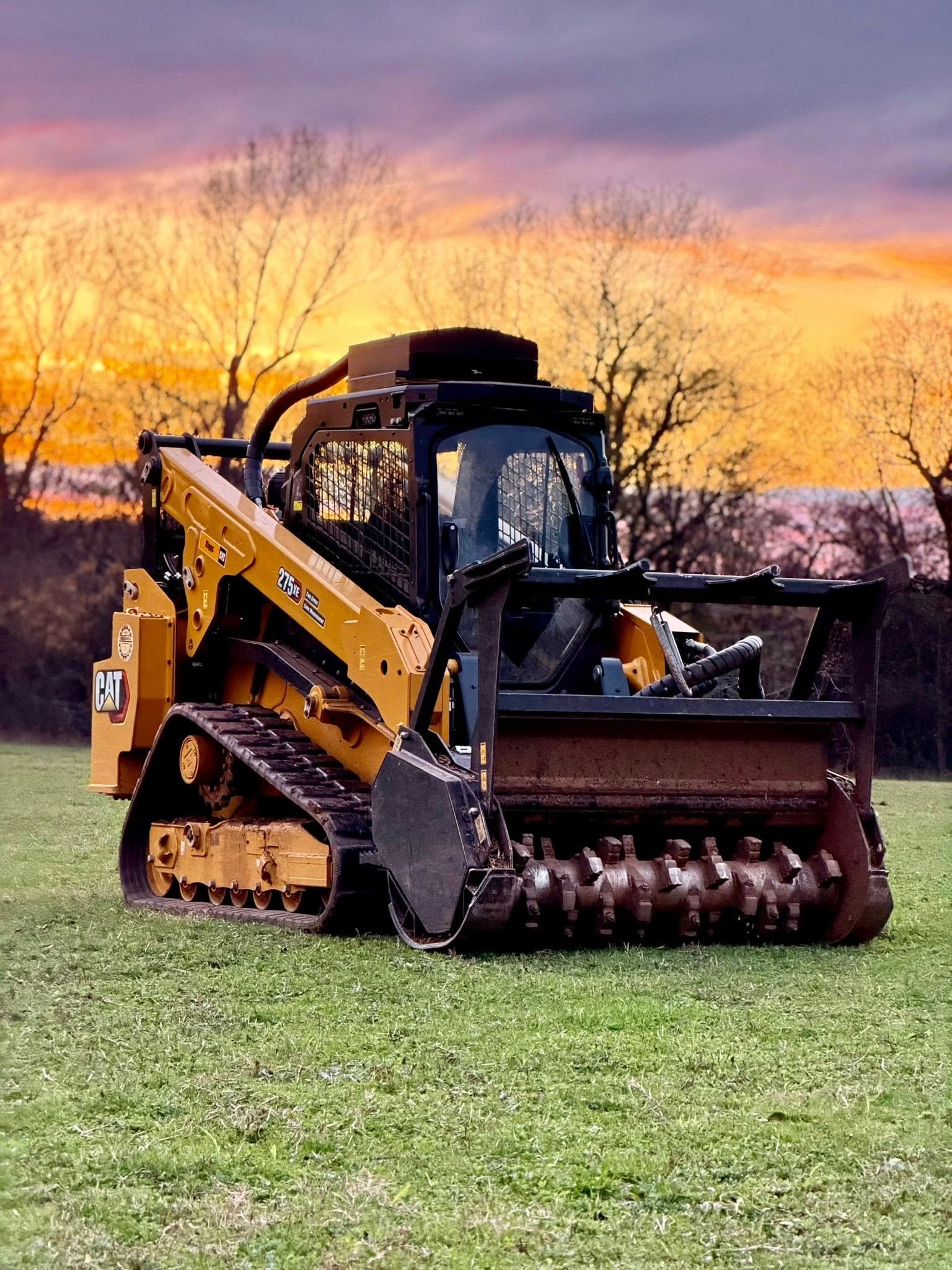 Yellow and black track loader with mulching head on a grassy field, under a sunset sky.