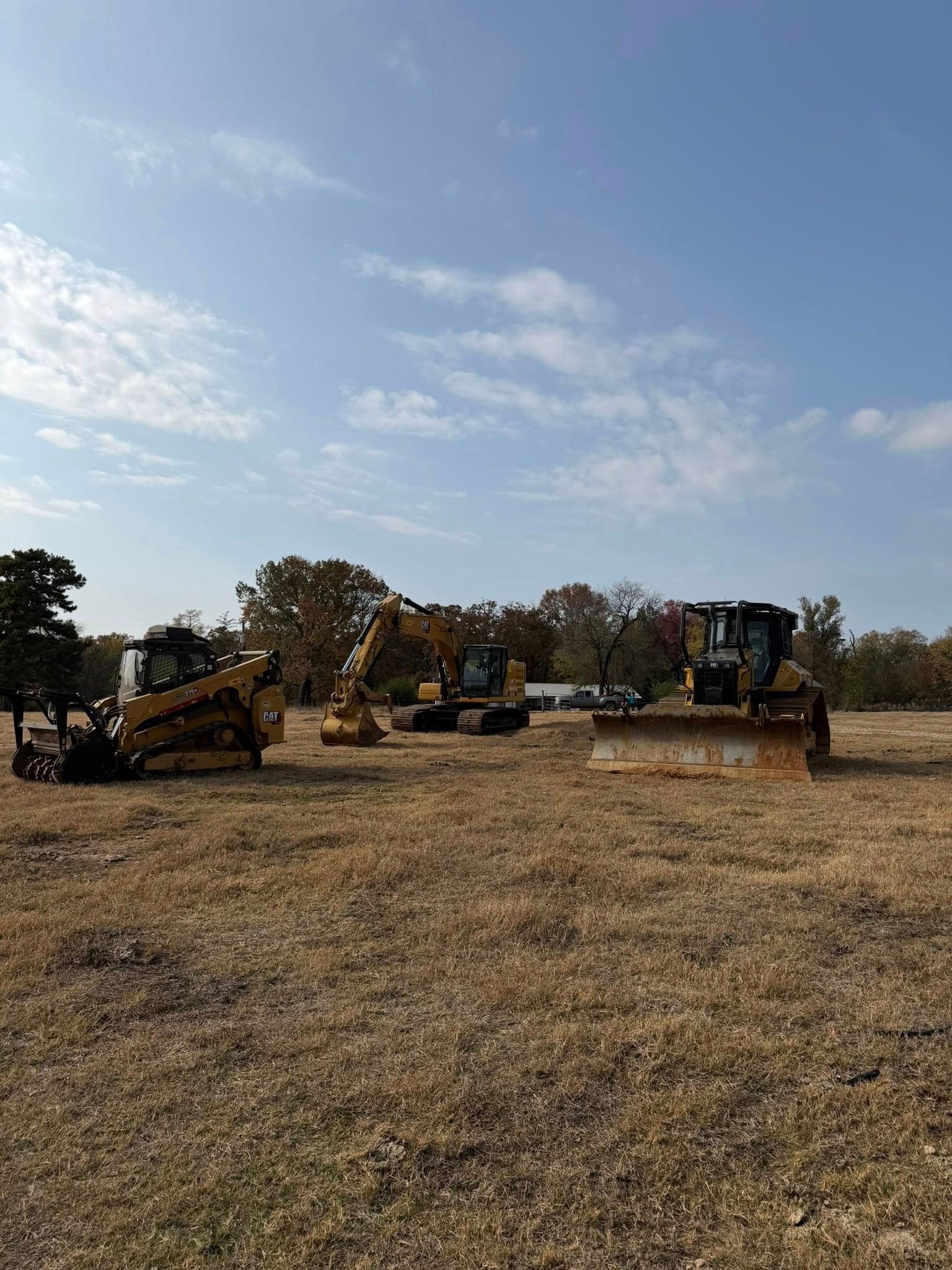 Three yellow construction vehicles parked in a dry, grassy field under a blue sky with clouds.