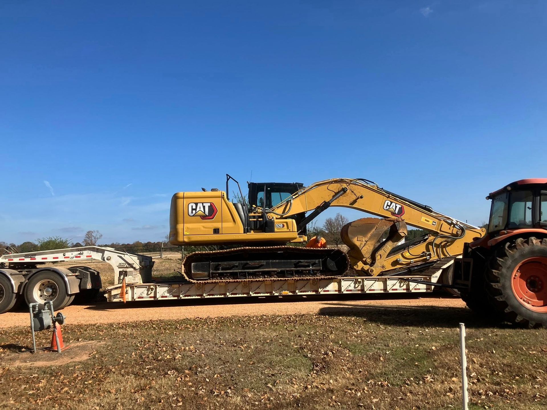 Yellow excavator on a flatbed trailer, pulled by an orange tractor, on a dirt road under a blue sky.