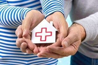 Dos manos sosteniendo un cartel con forma de casa con una cruz médica roja, que simboliza la atención médica en el hogar.