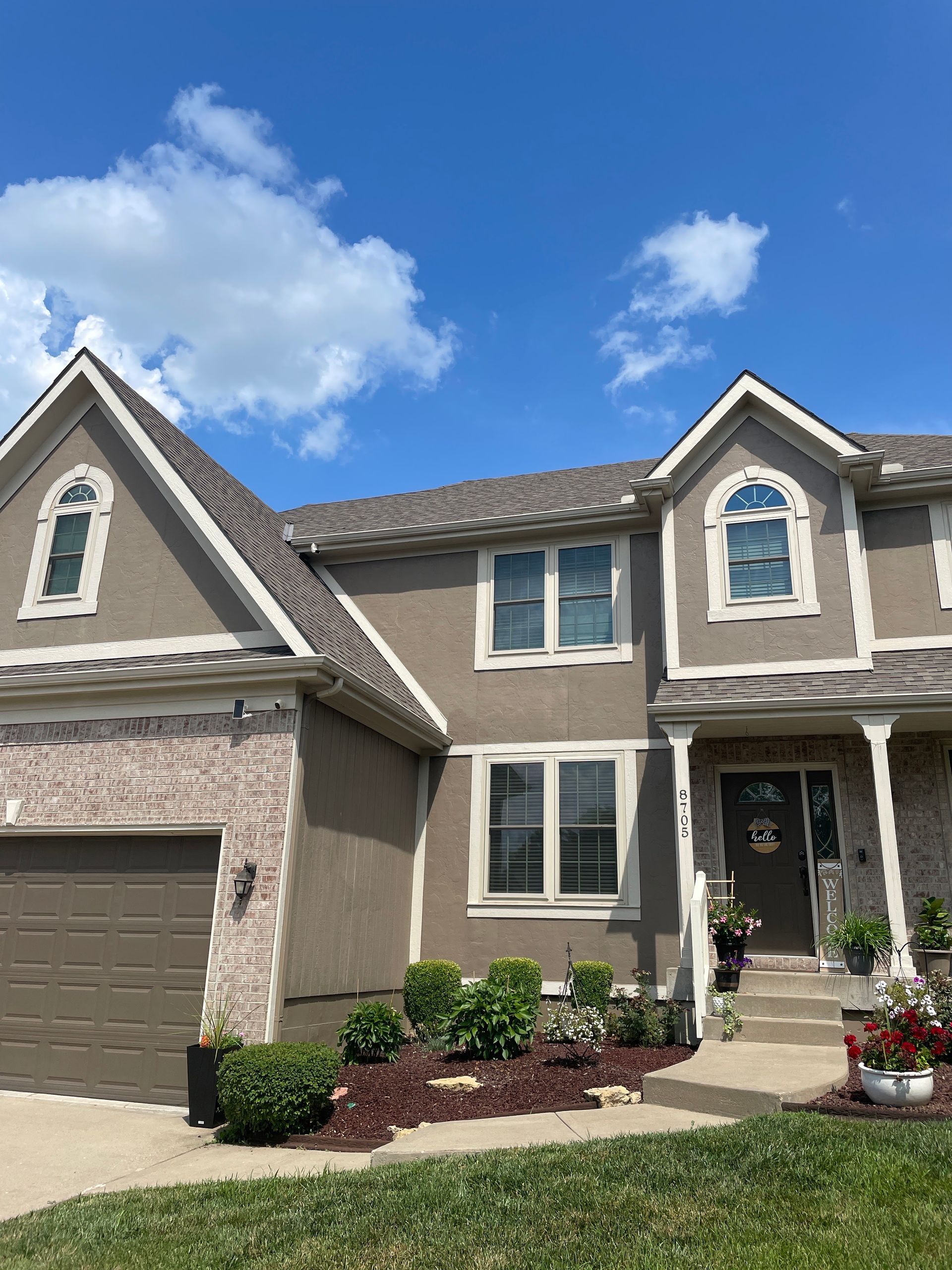 Suburban two-story tan house with gray roof, white trim, front porch, and attached garage under a blue sky