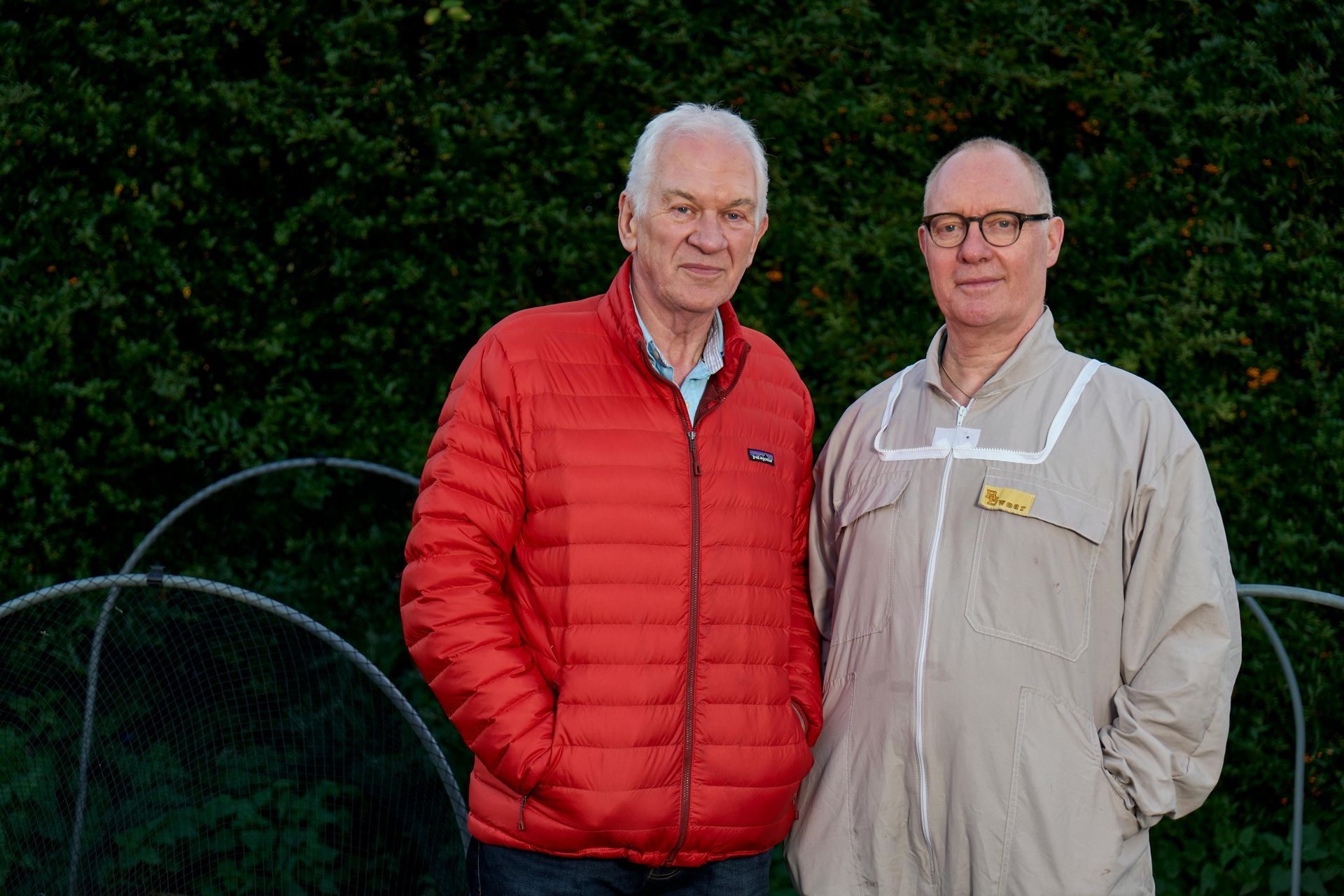 Two men are standing next to each other in front of a greenhouse.