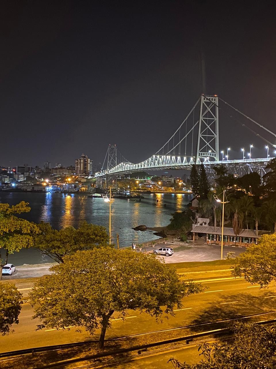À noite, a Ponte Hercílio Luz, iluminada, atravessa as águas, conectando o horizonte da cidade sob um céu escuro.