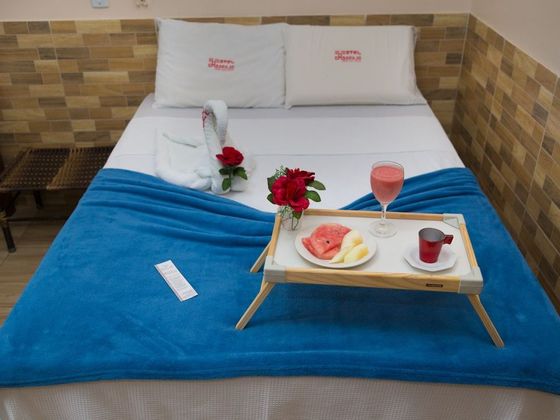 Uma cama de hotel feita com lençóis brancos e uma manta azul, coberta com uma toalha em forma de cisne, uma rosa e uma bandeja de café da manhã com frutas.