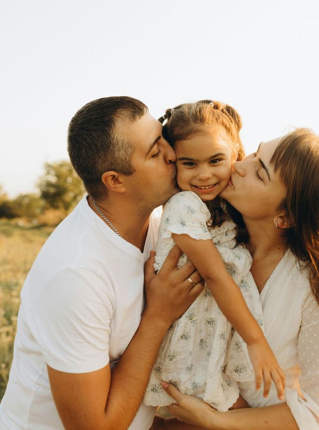 A family sharing a close kiss outdoors in warm sunlight, with a child smiling between two adults.