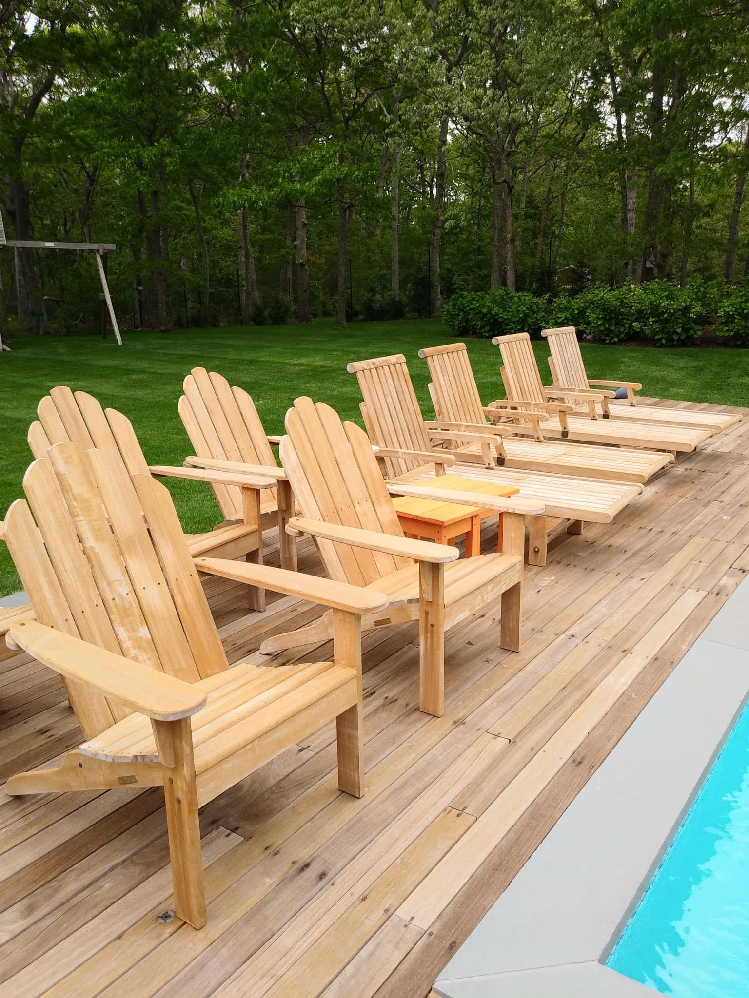 Wooden Adirondack chairs on a deck next to a pool, overlooking a green lawn and trees.
